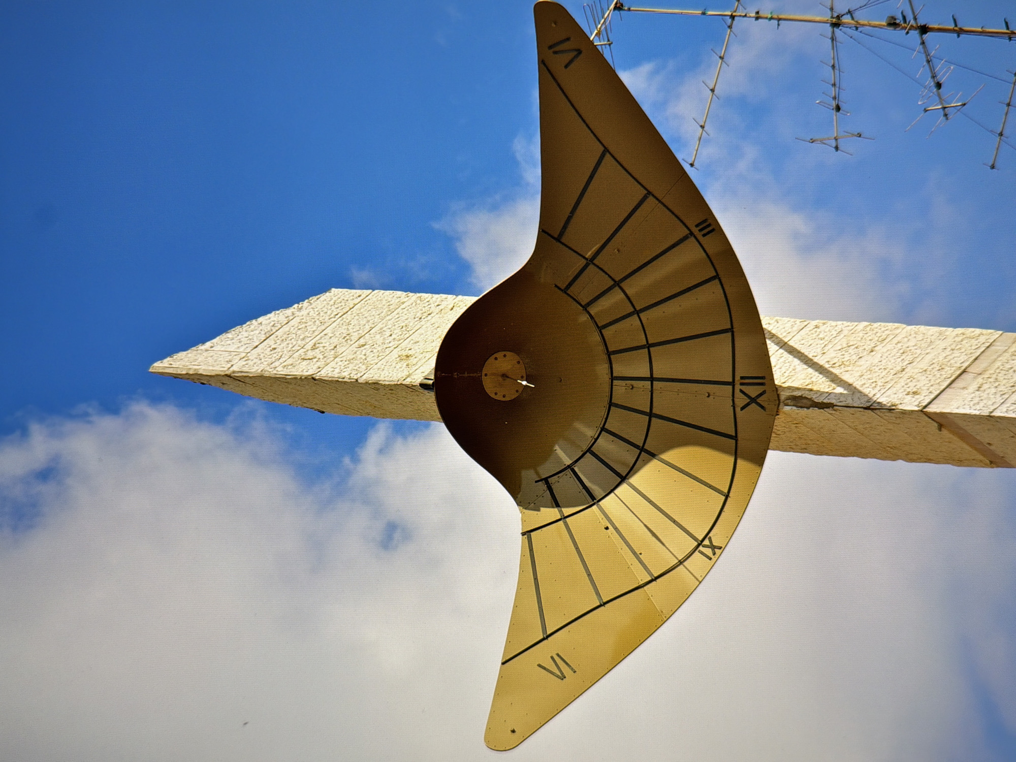 An outdoor sundial, constructed of a golden-yellow metal, is mounted on a tall, pale stone obelisk against a bright blue sky with scattered white clouds. The sundial features Roman numerals indicating hours, with "VI" visible on the left wing, "IX" on the lower part, and "XII" at the apex. A gnomon, the shadow-casting part of the sundial, is visible at the center of the device. The stone obelisk is textured with a rough, blocky pattern. In the background, to the right of the obelisk, a multi-tiered antenna structure is visible against the sky. The image does not contain any people. The scene appears to be in an open, outdoor location.