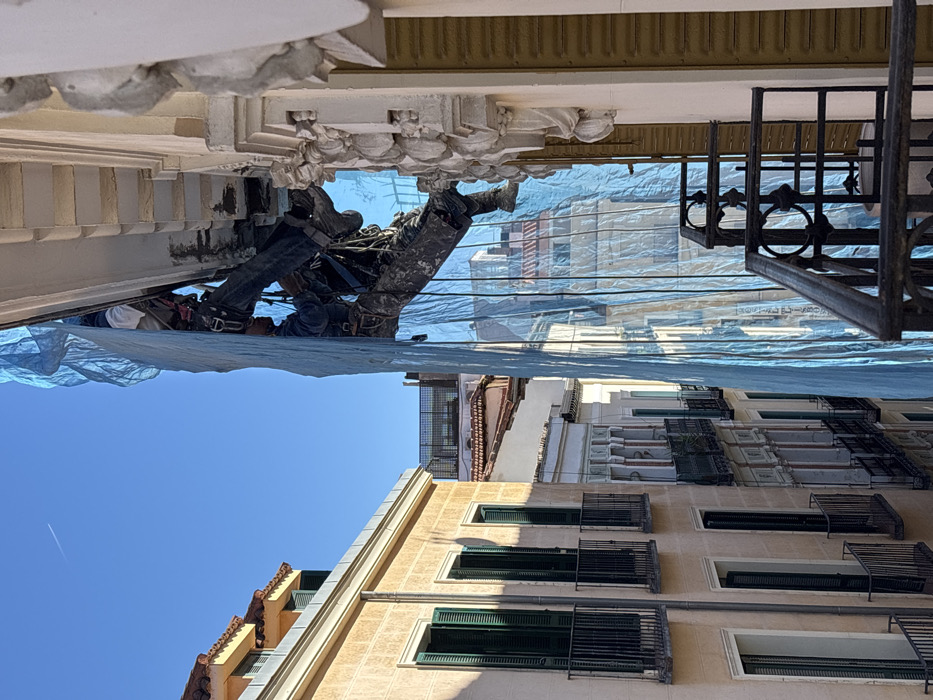 The image captures a construction worker suspended by ropes from the ornate cornice of a building facade in Madrid, Spain. The worker, dressed in dark work clothing and a safety harness, is partially obscured by a translucent blue plastic tarp or netting that hangs vertically from the building. Visible sections of the cornice show signs of repair or deterioration, with exposed darker material underneath the light-colored, sculpted architectural elements. The perspective is from a nearby balcony, indicated by a black wrought-iron railing in the immediate foreground. To the left, a series of multi-story buildings with beige facades, dark green louvered shutters, and black balcony railings line what appears to be a narrow street or alley. The sky is clear blue with a faint airplane contrail, indicating bright daylight conditions.