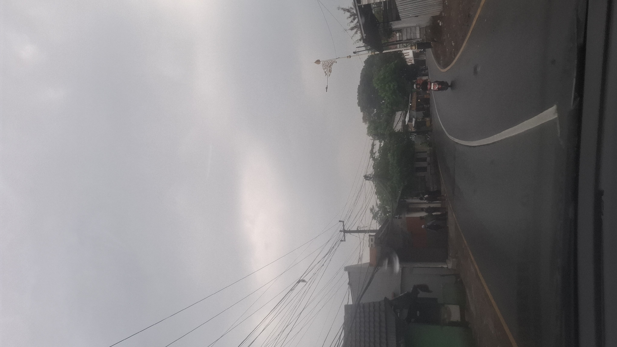The image depicts a street scene in Cilaku, Indonesia, under an overcast sky. A paved road curves through a residential area, with a white line marking its center. On the left side of the road, a row of houses with tiled roofs and some fences are visible, along with utility poles and a dense network of overhead wires. A person is standing near the entrance of one of the houses. On the right side of the road, more houses and trees line the street, and a decorative lamppost with an ornamental top stands near the road. A motorcycle is being ridden down the road, approaching the viewer. The ground appears wet, suggesting recent rain.