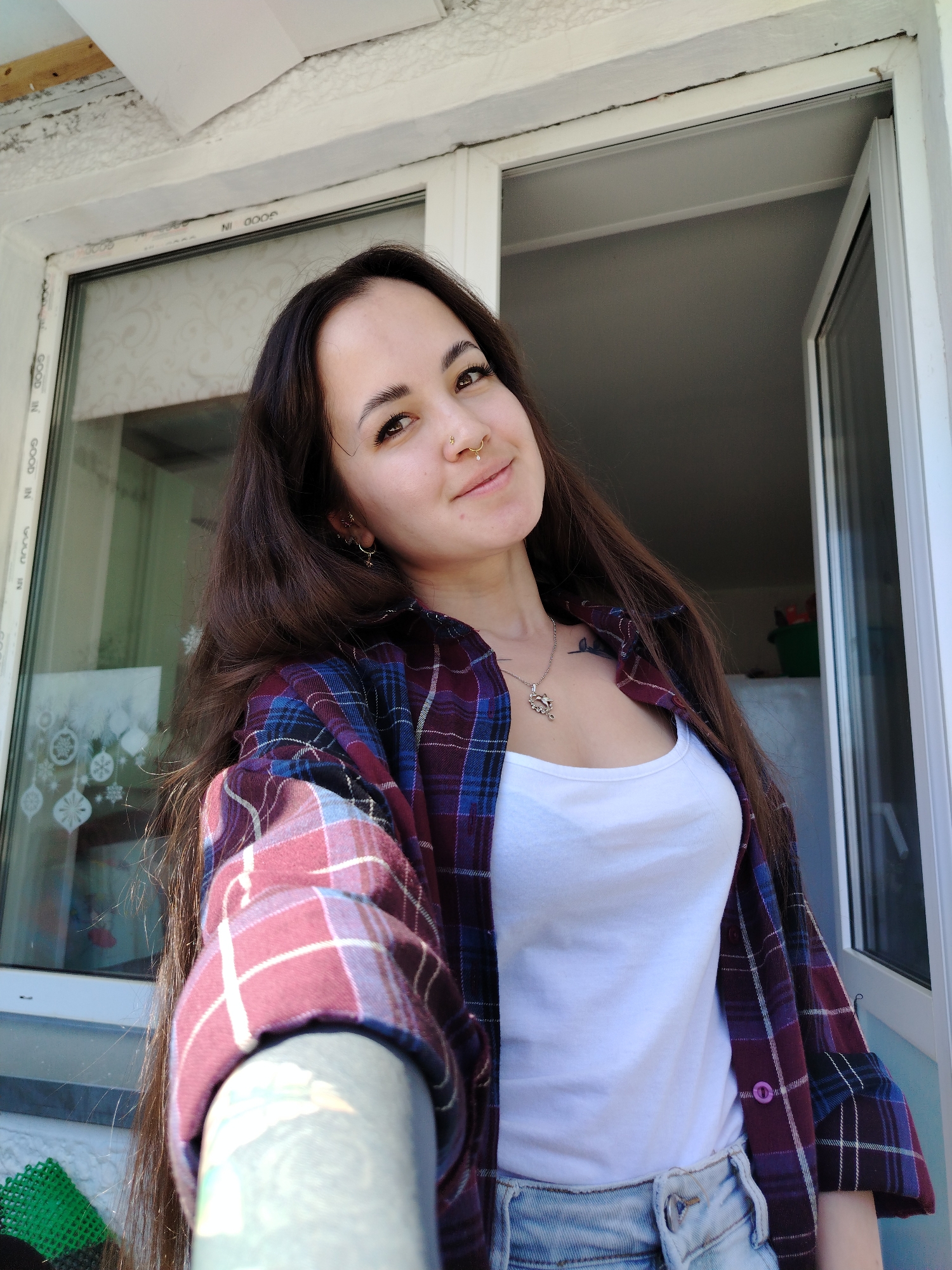 A young woman stands indoors near a window. She has long dark hair, and is wearing a white tank top, a red and blue plaid shirt, and denim shorts. She has piercings in her nose and ears, and a necklace. A window is behind her, and another window is slightly opened to her right. The location is Noviy, Russia.