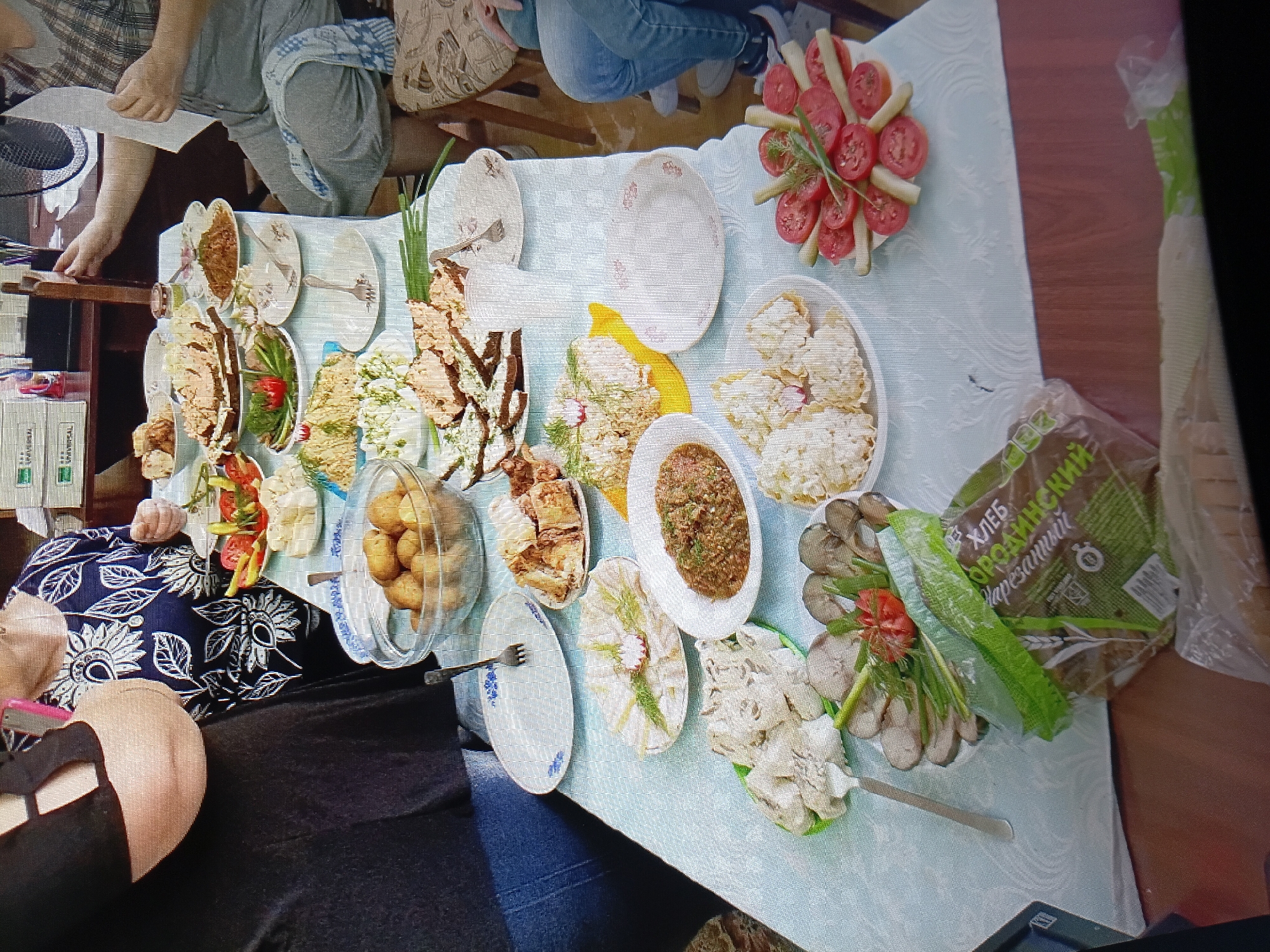 The image depicts a table laden with a variety of food dishes, suggesting a meal or gathering. In the foreground, a loaf of dark bread in plastic packaging is visible, with text in Cyrillic. Various plates contain sliced meats, vegetables like tomatoes and pickles, and what appears to be salad or a mashed dish. There are also bowls of potatoes and other prepared foods. The table is covered with a light blue, patterned tablecloth. Several people are partially visible in the background, seated around the table, with one person in a black top and another in a blue and white floral patterned blouse. The environment suggests an indoor setting, possibly a dining room or kitchen, with a glimpse of office-like supplies in the upper background and a fan. The image quality is somewhat low, with visible pixelation. Based on the provided information, it is not possible to determine the specific location in the city of Tiraspol, Moldova.