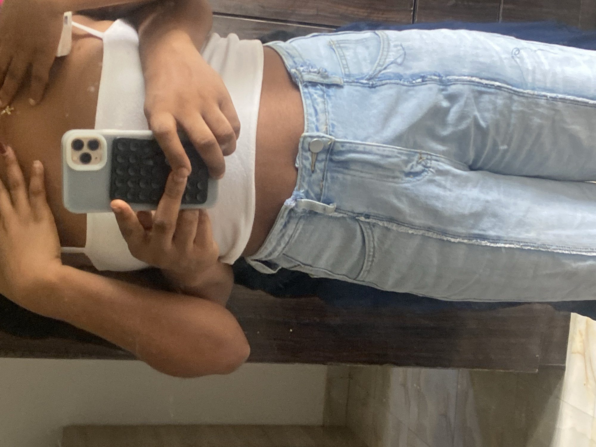 A person is lying on a dark brown surface, wearing light blue jeans and a white tank top. They are holding a gray iPhone in their right hand, taking a selfie. The background includes a marble-like surface. The specific location within Abuja, Nigeria is not discernible from the image.