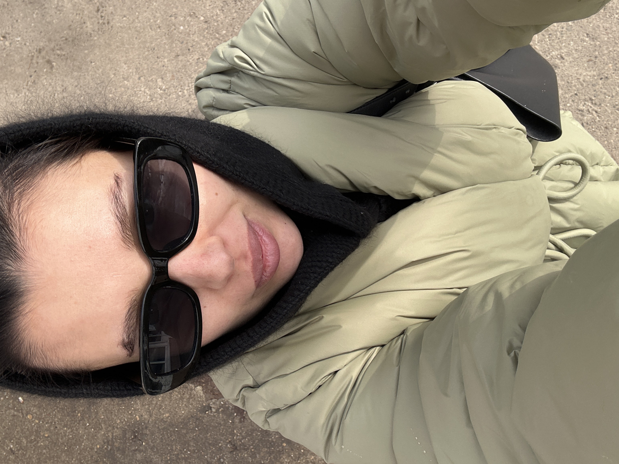 The image displays a high-angle selfie of an individual in Chaikovsky, Russia. The subject is an adult female, positioned with her head slightly tilted forward, looking directly at the camera. She wears black, rectangular-framed sunglasses that obscure her eyes. Her dark hair is mostly covered by a black, textured hood or balaclava-style head covering. Her face shows a visible forehead, dark eyebrows, and full lips with a light pink tint. She is dressed in a puffy, light olive-green or sage-colored winter jacket, which appears to have a slightly glossy finish and horizontal segmentation. A drawstring is visible at the lower front of the jacket. A black strap and a portion of a black bag are visible on her right side, partially tucked under the jacket. The background consists of a light brown or grey granular surface, consistent with ground pavement or a similar outdoor surface, visible in the upper and lower portions of the frame. Reflections in the sunglasses indicate an outdoor environment with ambient light and a distant structure, possibly a window.