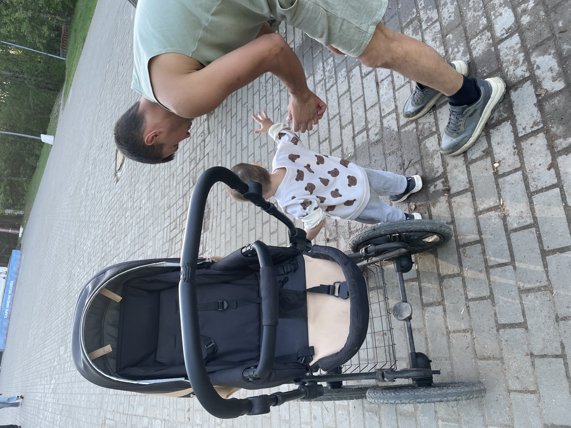 A man and a young child interact beside a stroller on a paved brick surface. The man, wearing a light green t-shirt and shorts, leans down, holding the child's hand. The child, dressed in a white sweatshirt with brown bear designs and gray pants, reaches towards the man. A black stroller with a beige interior sits nearby. The location is a park, specifically named "Парк имени Чехова", in the city of Perm, Russia. The scene is well-lit, suggesting daytime.