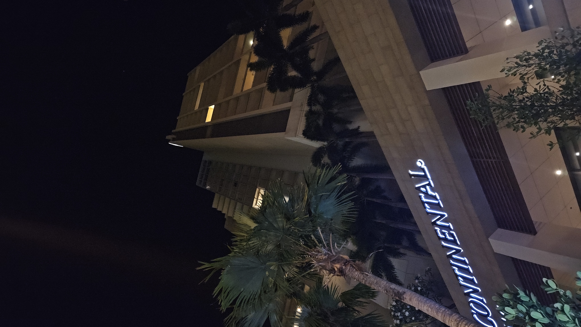 A nighttime shot from below, looking up at a modern building with the word "CONTINENTAL" vertically displayed in blue neon lights. The building is likely a hotel, considering the word displayed. Several palm trees partially obscure the view of the building. The building is light-colored, possibly beige or light gray. It has a modern architectural design with sharp angles and rectangular windows. The sky is dark, indicating it's nighttime. The location is Nha Trang, Vietnam, implied by the presence of the Continental Hotel in the city.