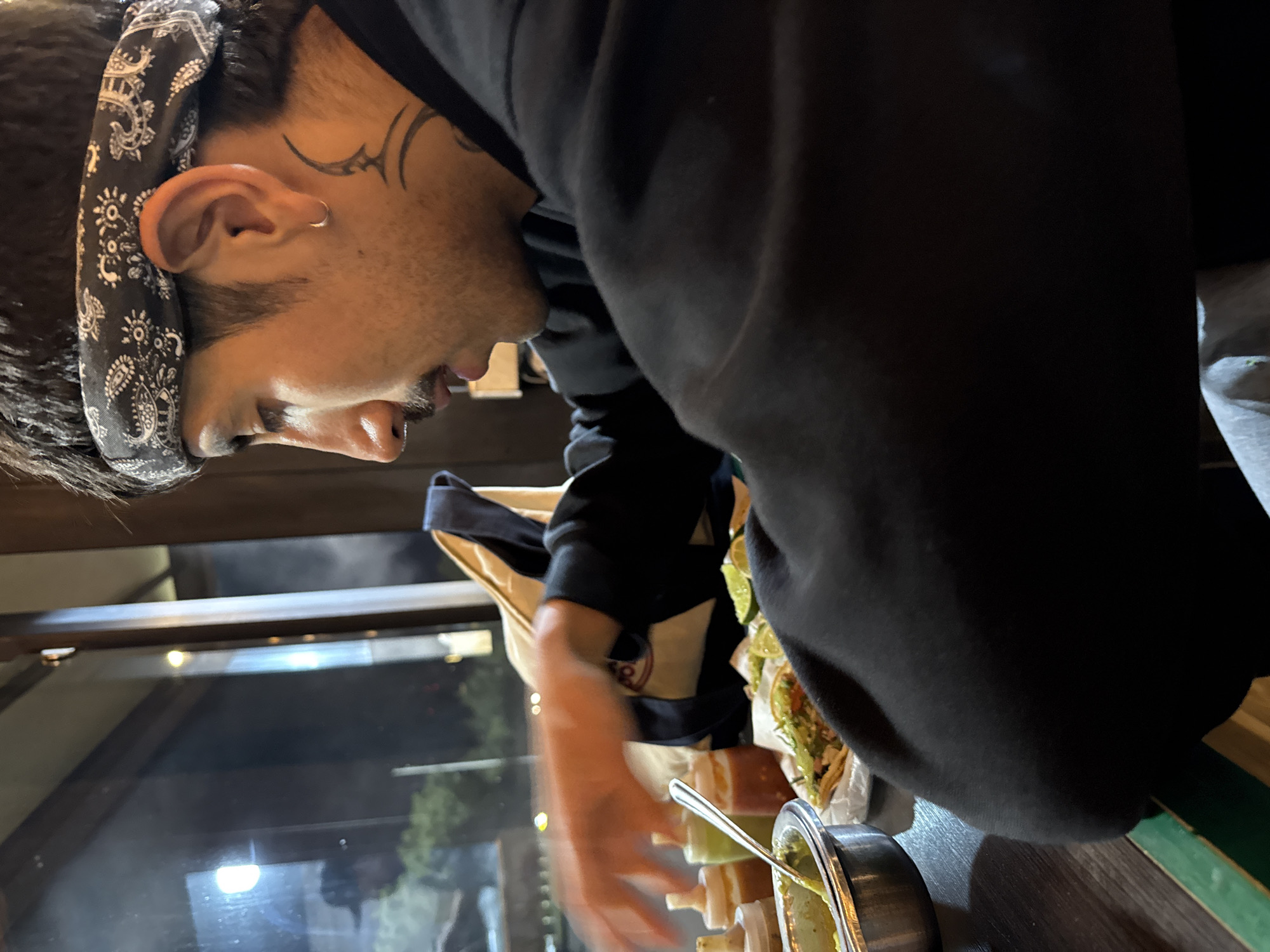 An indoor scene depicts a male individual seated at a dark table in what appears to be a restaurant in San Bernardino Tlaxcalancingo, Mexico, during nighttime hours. The individual, seen in profile from the chest up, is positioned facing right. He wears a black and white paisley-patterned bandana, a small silver hoop earring in his left ear, and a black tribal-style tattoo extending down his left neck. His facial hair includes a light mustache and goatee. He is dressed in a dark, possibly black, long-sleeved garment. His left hand is blurred, indicative of motion, as it reaches towards a meal laid out on the table.
The table features a white paper-lined plate holding several tacos or similar food items topped with green and red ingredients, accompanied by lime wedges. To the left, a silver bowl contains a green substance, likely guacamole or a green salsa, with a spoon resting inside. Behind this, four plastic squeeze bottles of various sauces—red, light green, orange, and off-white—are arranged. A large window to the left reveals a dark exterior with blurry reflections of a person wearing a hat and streetlights. A light-colored bag is partially visible behind the subject near the window. The overall environment suggests a casual dining setting.