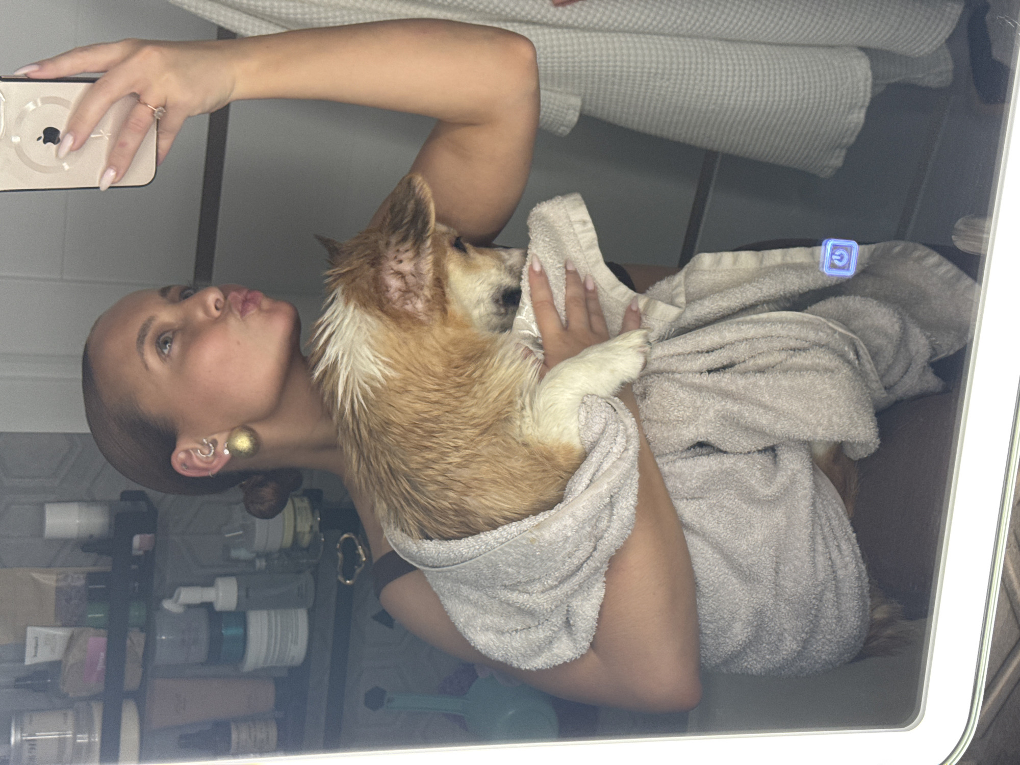 A woman stands in a bathroom, holding a small dog wrapped in a gray towel. She is taking a selfie with a gold-colored iPhone, puckering her lips towards the camera. She has tan skin, short hair in a bun, and wears multiple earrings. The bathroom has a mirrored cabinet containing toiletries. A second gray towel hangs above. The location is Chaikovsky, Russia.