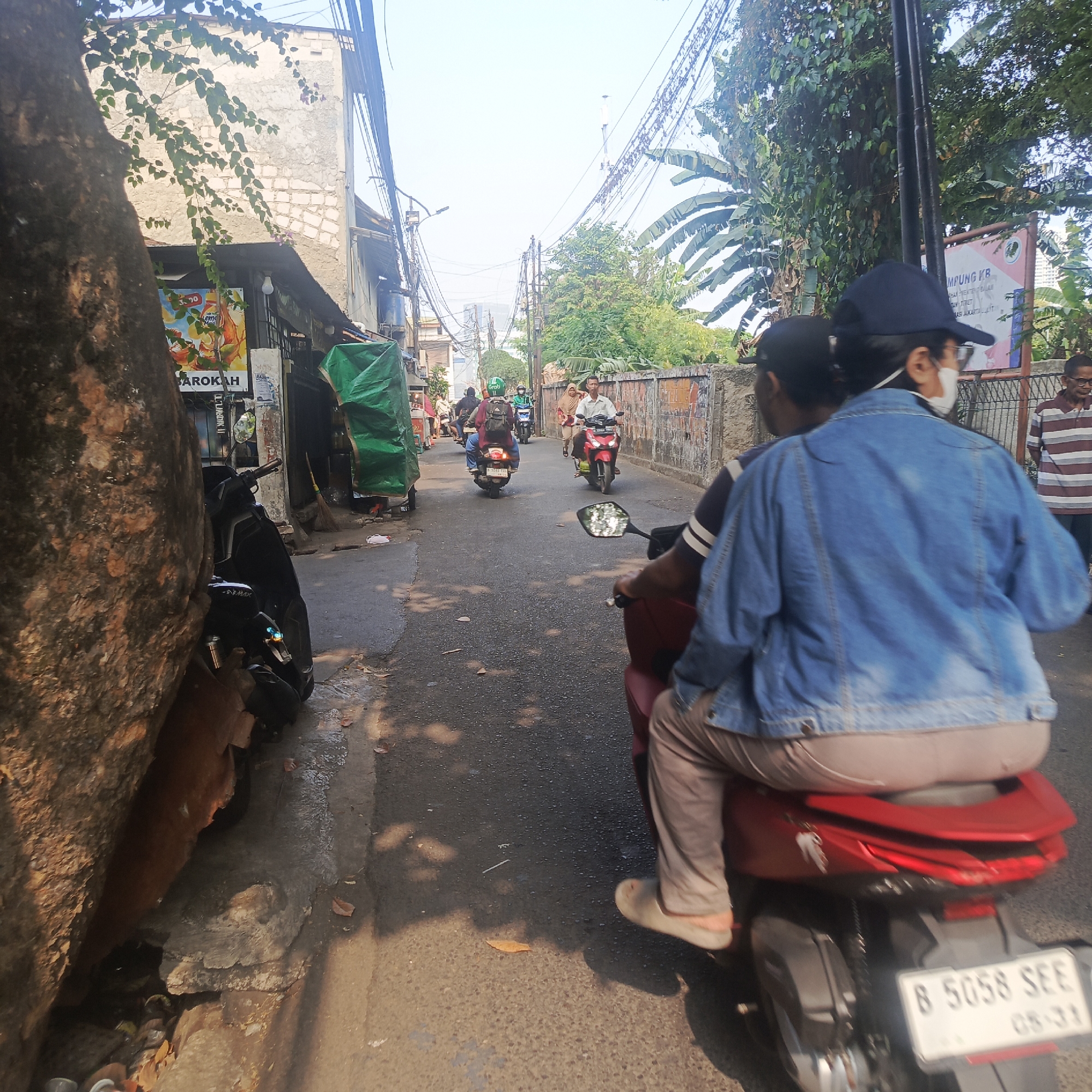 The image shows a street scene in South Jakarta, Indonesia. A motorbike, with two people on it, is in the foreground, traveling away from the viewer. The person in the back is wearing a denim jacket and a mask. In the middle ground, other motorbikes are seen traveling in the same direction. The street is lined with buildings and trees, and there are many electrical wires overhead. A large tree trunk is visible on the left side of the image. The scene appears to be a typical urban environment in Indonesia, with motorbikes being a common mode of transportation.
