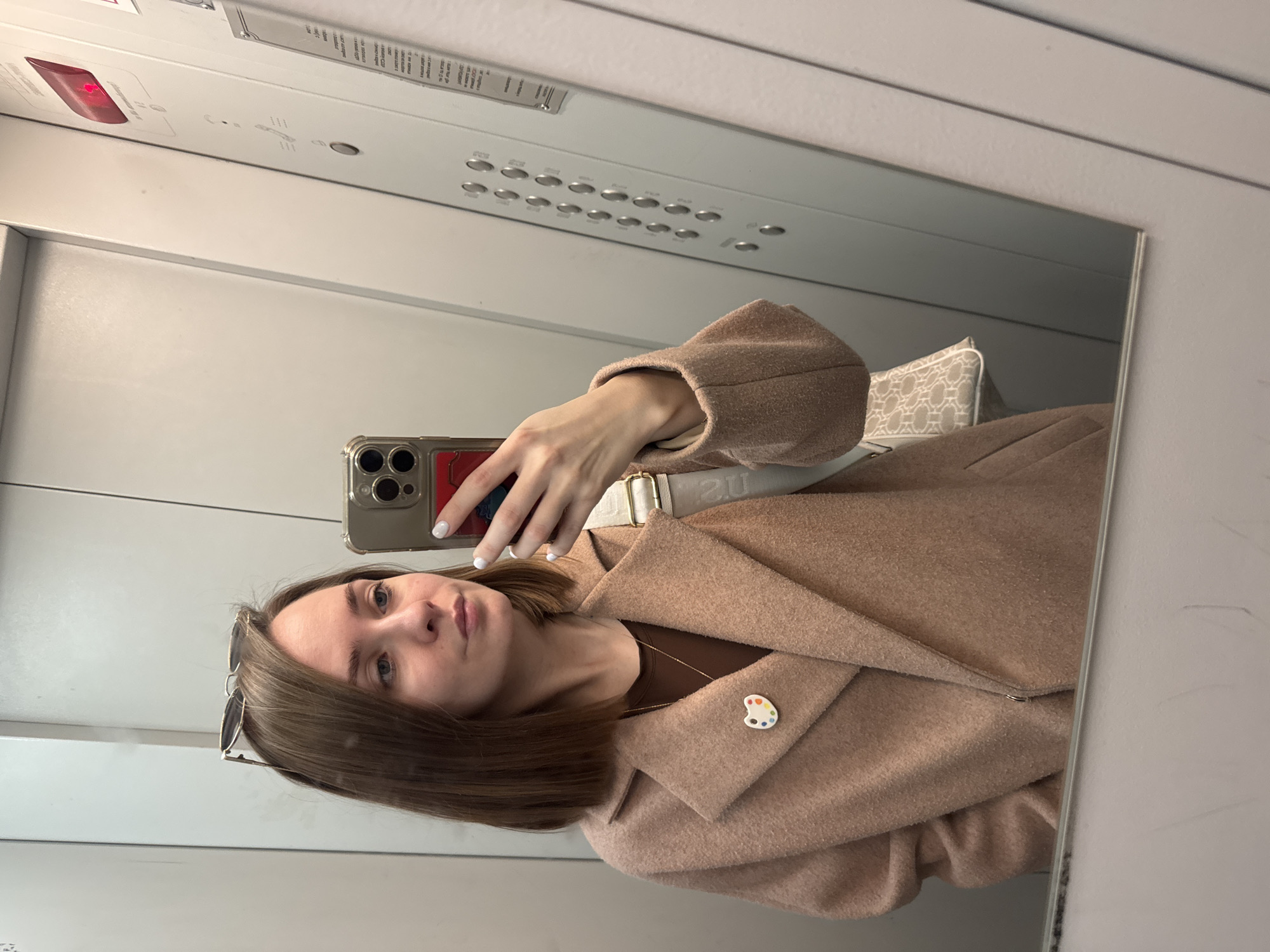 The image is a selfie taken in what appears to be an elevator. A young woman with light brown hair is holding a smartphone, a newer model possibly an iPhone based on the camera design, to capture her reflection in a mirror. She is wearing a light brown coat with a colorful artist's palette pin on the lapel, visible against a dark brown top with a gold necklace. Sunglasses are perched in her hair. The elevator's control panel is visible above her reflection. The environment is clean and sterile, suggesting a modern elevator. Based on the metadata for location information, the image was taken in Perm, Russia.