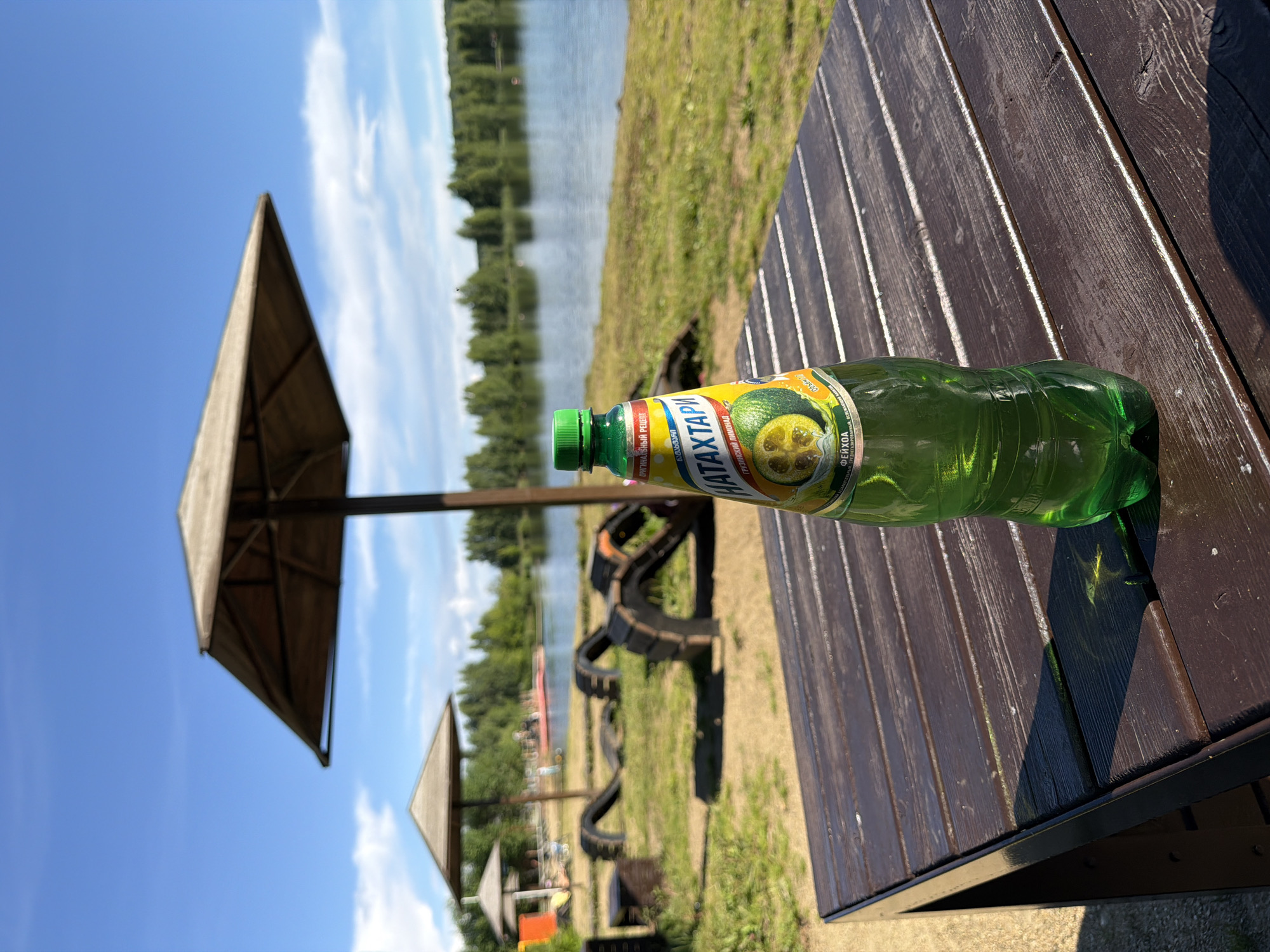 A green plastic bottle of beverage rests on a dark wooden picnic table, positioned diagonally across the frame. The bottle is partially filled, with its label featuring a picture of a feijoa and the text "НАТАХТАРИ" in large, bold letters. The scene is set outdoors, with a body of water visible in the background, bordered by a grassy bank and trees. A wooden umbrella shelter stands to the left of the bottle, casting a shadow. The sky is blue with scattered white clouds, suggesting a sunny day. The overall impression is a leisurely outdoor setting, possibly a park or recreational area by a lake. The sunlight indicates it is likely daytime. There are no people clearly visible in the image, but the presence of the picnic table and umbrella suggests a place intended for human use and relaxation. The bottle and the picnic table are the primary objects in focus.