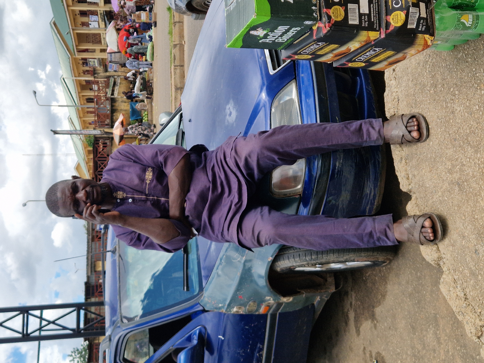 A dark-skinned man with short hair and a beard is sitting on the hood of a blue car, talking on a dark mobile phone. He is wearing a purple traditional African garment and brown sandals. His arms are crossed. The car appears to be parked on a dirt and paved surface. To the right of the car, there are stacks of boxes containing beverages, including "Action Bitters" and "CREMICA". In the background, there is a street scene with buildings, market stalls, umbrellas, and other people. The sky is cloudy.