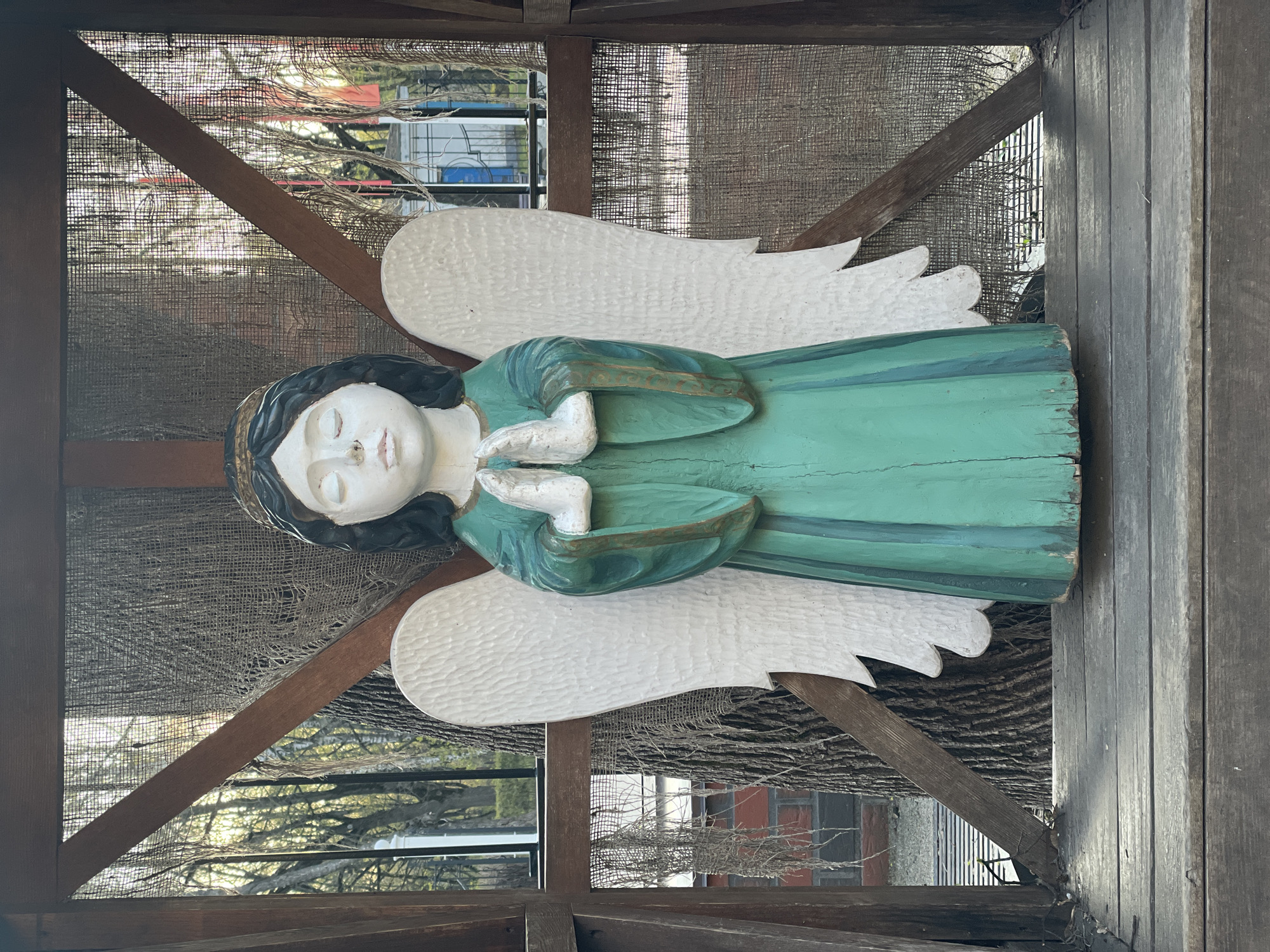 A medium shot of a wooden, painted angel sculpture. The angel is mostly green with white wings, white hands, and a white face. It appears to be indoors, with a wooden framework surrounding it, and a scrim-like material behind it. Beyond the scrim, trees and buildings are visible. There is no evidence to indicate that this image was taken in Khimki, Russia.