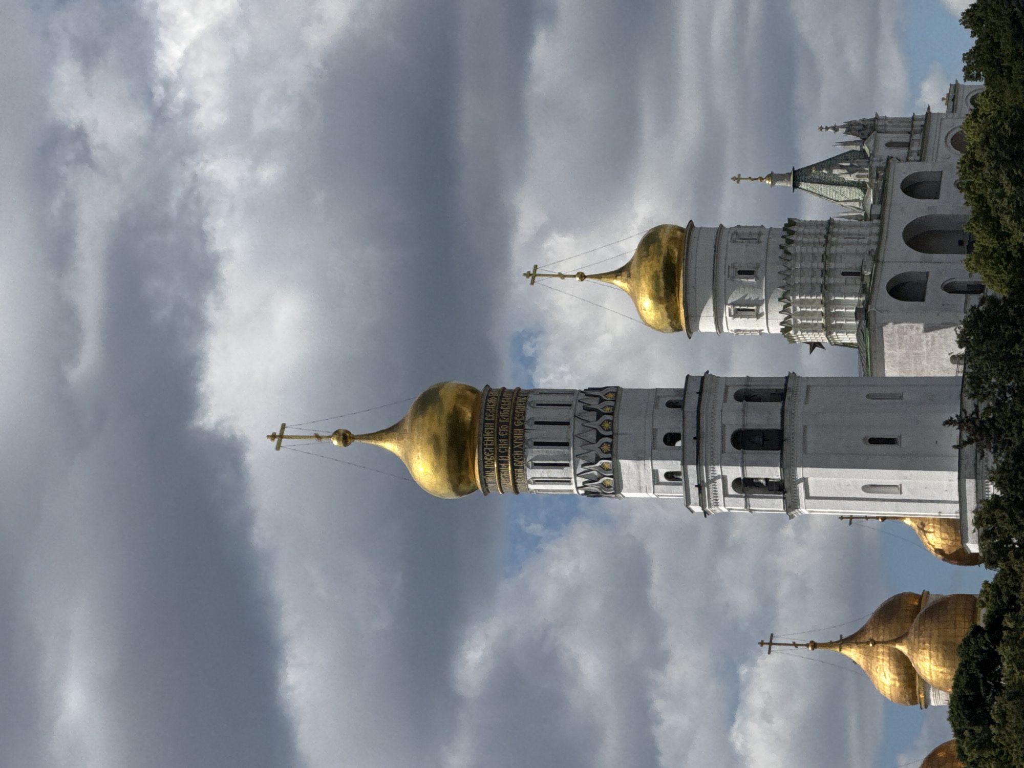The image shows a close-up view of the Ivan the Great Bell Tower and surrounding cathedrals within the Moscow Kremlin, Russia. The bell tower, a tall, white, multi-tiered structure topped with a golden dome and cross, dominates the center of the frame. To its right, another white building with a smaller golden dome and cross is visible, along with parts of other structures. Golden domes of other churches are also visible on the left and right edges of the image, peeking above a dense line of dark green trees. The sky is filled with dramatic, dark gray clouds, with patches of lighter sky visible in between. The scene is one of architectural grandeur set against a turbulent sky, with no people or animals present.