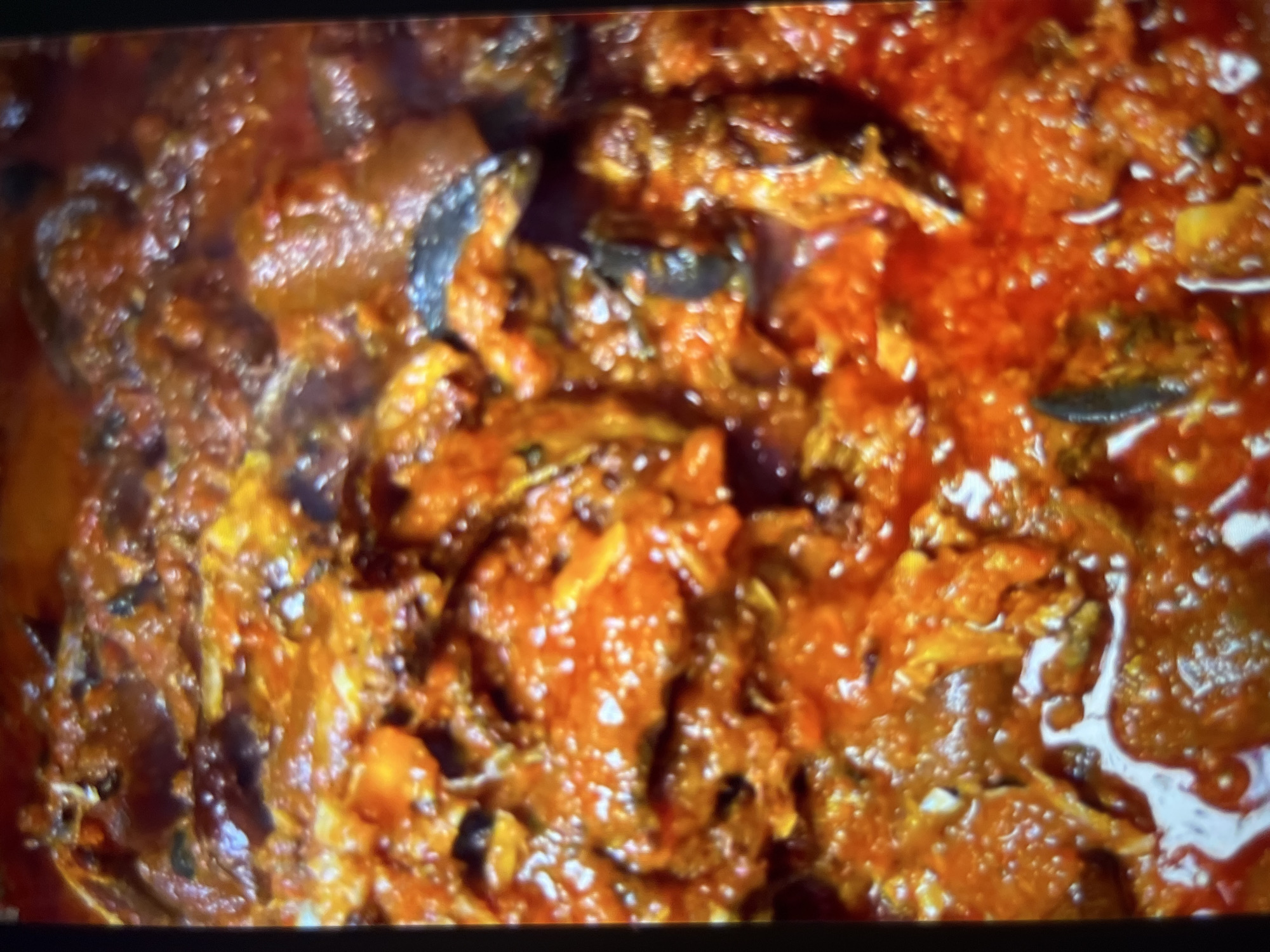 The image displays a close-up, overhead view of a dish of stew, likely a traditional Nigerian cuisine, with a focus on the textures and colors of the ingredients. The stew is a rich, reddish-orange hue, characteristic of tomato-based stews, and appears to be a hearty mixture of fish, possibly dried or smoked, and pieces of meat or offal. The surface of the stew is uneven, with some areas appearing smoother and more liquid, while others show a thicker, more textured consistency. There are visible chunks of ingredients, some darker and some lighter, interspersed throughout the sauce. The overall impression is one of a flavorful and robust dish. The image does not provide any information about its specific location within the city of Abuja, Nigeria, nor does it depict any people, actions, or environmental context. It is solely focused on the presentation of the food.