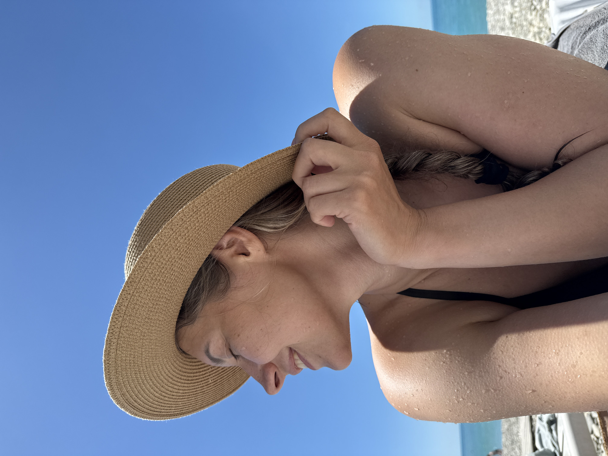 An outdoor, close-up, head-and-shoulders shot shows a young woman wearing a straw hat and a black bikini top. She is looking to her left with her eyes closed and a slight smile. Her hand is raised, holding the brim of her hat. Her skin is wet with droplets of water visible on her arm and shoulder. The background is a clear blue sky and the sea in the distance, suggesting a beach location. The image is taken in Nebug, Russia.