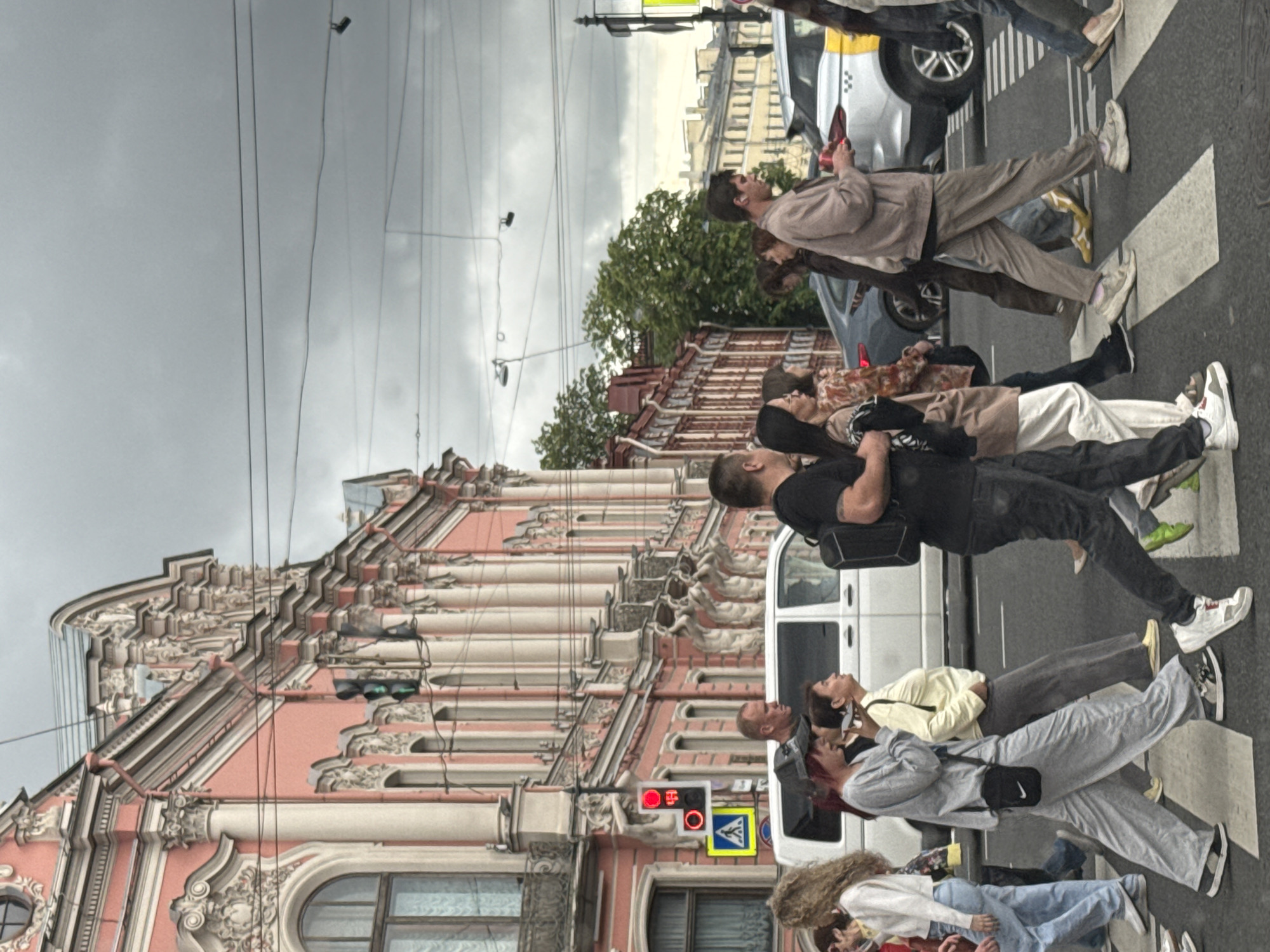 The image captures a street scene in Saint Petersburg, Russia, on a cloudy day. A pedestrian crossing is occupied by several individuals walking in various directions. In the background, a white van is parked, and a silver car with yellow detailing is in motion. Dominating the left side of the frame is a multi-story building with ornate pink and cream-colored architecture, characterized by columns, decorative carvings, and arched windows. Traffic lights, one red and one green, are visible, along with a pedestrian crossing sign. The sky is overcast, and power lines crisscross the upper portion of the image. The overall impression is a typical urban environment with an emphasis on architectural details and everyday human activity.