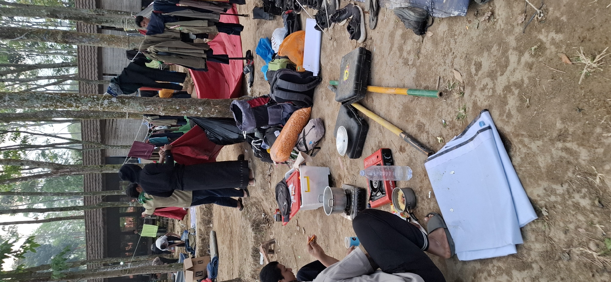The image depicts an outdoor scene likely in Puntukrejo, Indonesia, characterized by a collection of camping or temporary living supplies amidst trees and a low-lying structure. In the foreground, a person wearing black pants and gray slippers is seated and appears to be eating. Various cooking items are scattered around, including a portable gas stove with a pot on it, a small pot with food, and a bottle of water. Other objects visible include several backpacks, a large orange bag filled with what appears to be snacks, a white cooler, and a folded light blue tarp. In the mid-ground and background, more people are present, some tending to laundry hung on a line between trees. Clothing is visible on the line, as are larger tarpaulins, one red and one blue, suggesting a temporary shelter setup. The environment is a dirt ground with sparse vegetation and numerous trees forming a canopy overhead, indicating a natural or semi-rural setting.