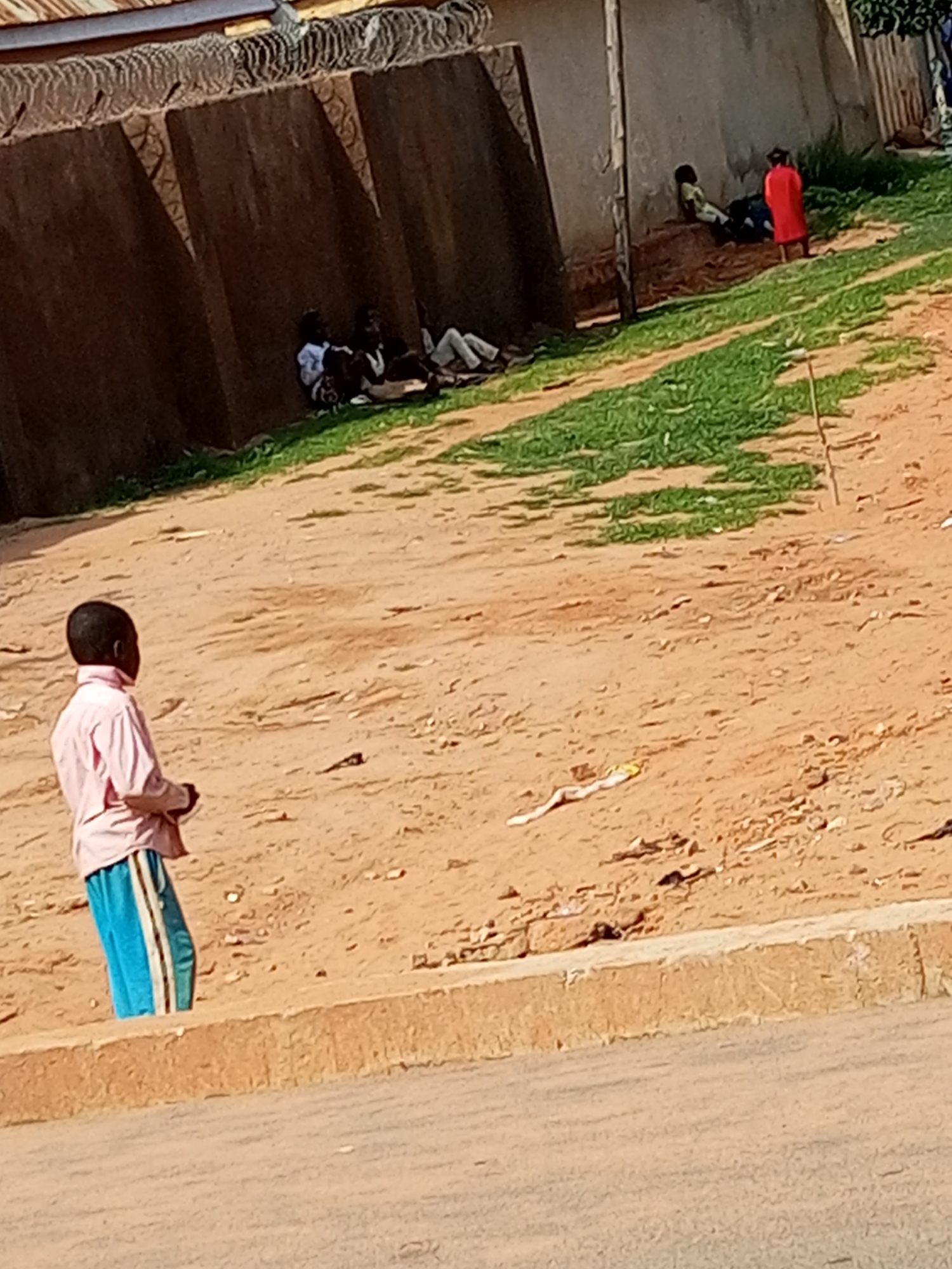 The image depicts an outdoor scene in Gombe, Nigeria, with several individuals present. In the foreground, a young person wearing a pink collared shirt and blue and white striped trousers stands facing away from the camera, looking towards the right side of the frame. The ground is primarily sandy with patches of green grass.
In the background, against a brown wall topped with barbed wire, a group of people are seated or lying down. One individual is wearing a bright red garment, while others are in darker clothing. The setting appears to be a public area, possibly a street or a yard adjacent to a building. The lighting suggests it is daytime, with sunlight casting shadows. There are no visible signs of text in the image. The overall impression is of a casual gathering or a moment of rest in a somewhat arid environment.