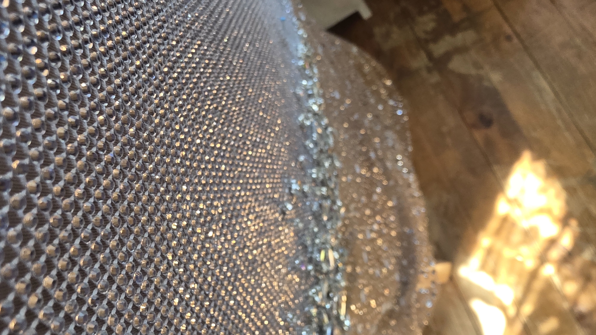 The image shows a close-up of a material covered in small, reflective, gem-like objects arranged in a grid pattern. The material is draped, with the edge visible and slightly blurred. To the right of the material is a wooden surface with a light source reflecting off of it. The light is bright and appears to be positioned at a low angle. There is no information available from the image to determine the location in the city of Blagoevgrad, Bulgaria. There are no people, subjects, or actions visible.