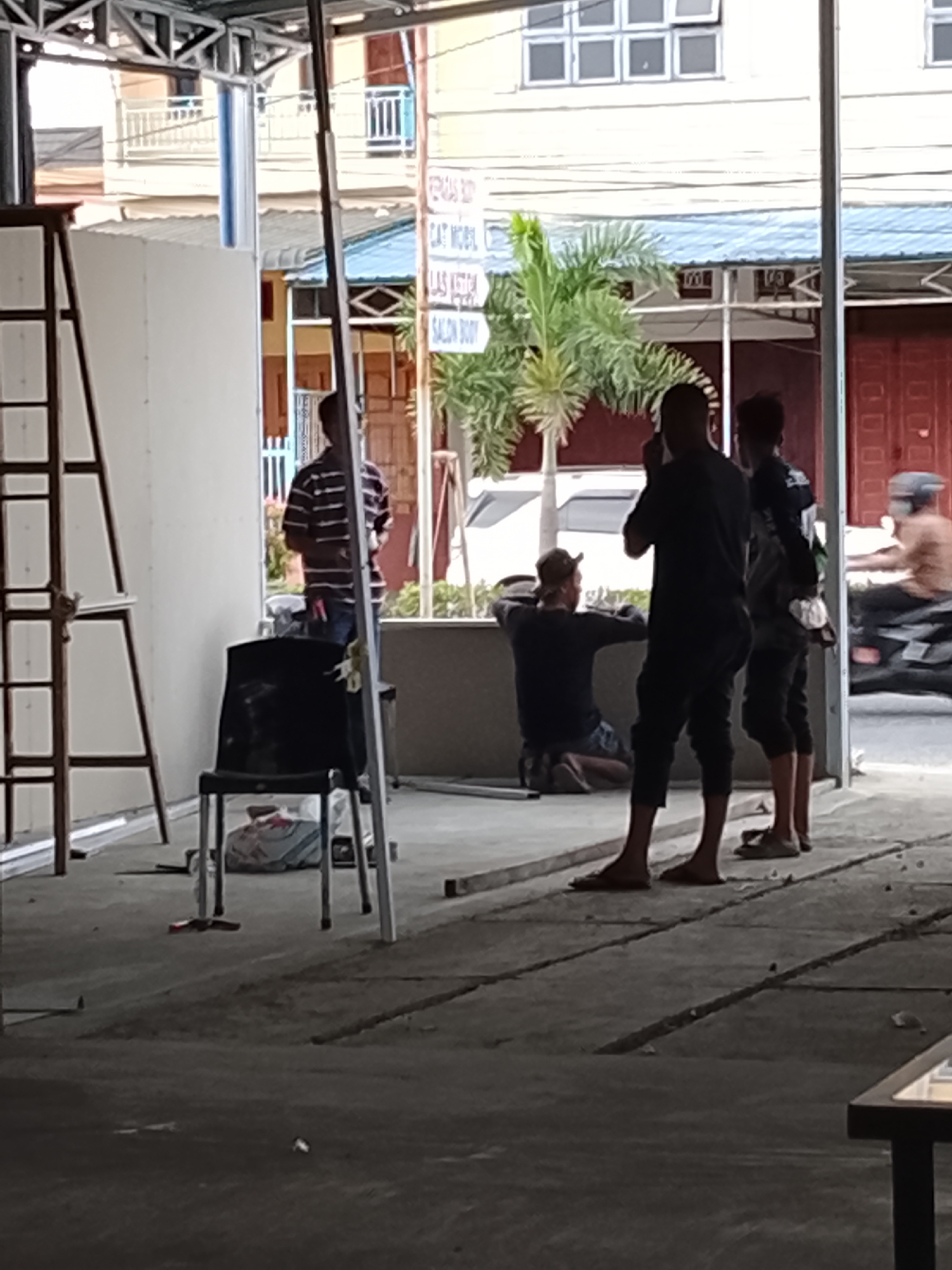 The image depicts an outdoor scene in Lhokseumawe, Indonesia. Several individuals are present, possibly engaged in construction or renovation work. A person in a striped shirt stands near a white wall and a ladder. Two men, one in a dark shirt and shorts, are standing looking to the right while another man is kneeling near a low wall or structure. A motorcycle rider is seen passing on the right. A chair sits on the concrete surface. In the background, buildings, a palm tree, and a sign are visible. The scene appears to be a street view with ongoing activity.