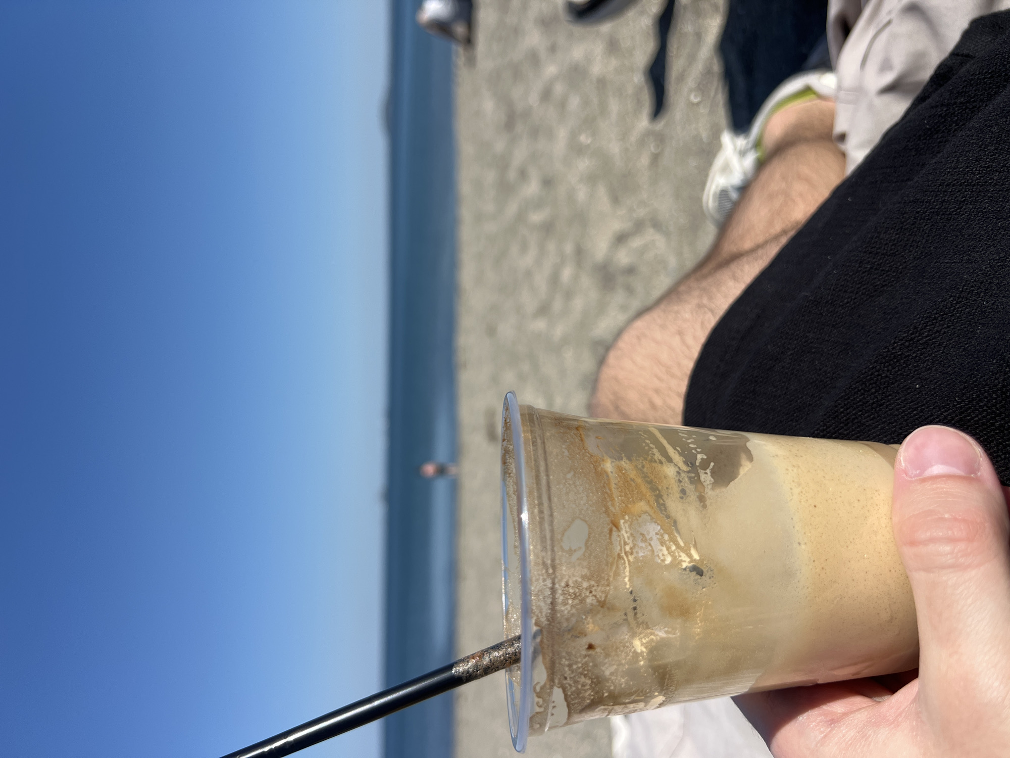 The photo is taken at Burgas Beach in Burgas, Bulgaria. It shows a partially consumed iced coffee drink in a clear plastic cup, held in someone's hand. A black straw is visible in the drink. In the background, there is a sandy beach and the Black Sea under a clear blue sky. A person is standing far away in the sea. Part of a person's legs and clothing are visible, suggesting the photo is taken from their perspective.