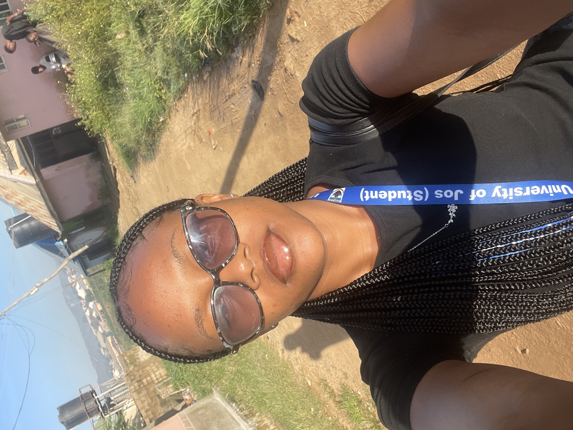 A young woman with braided hair and sunglasses takes a selfie outdoors. She is wearing a black t-shirt and a necklace. A blue lanyard hangs from her neck with the text "University of Jos (Student)" visible. The background shows a dirt path, overgrown grass, and buildings under a clear sky. The scene appears to be in a residential or semi-urban area, possibly Rafin Par, Nigeria, given the architecture and vegetation. There are other people in the background, but they are not the main focus of the image. The lighting suggests it is daytime.