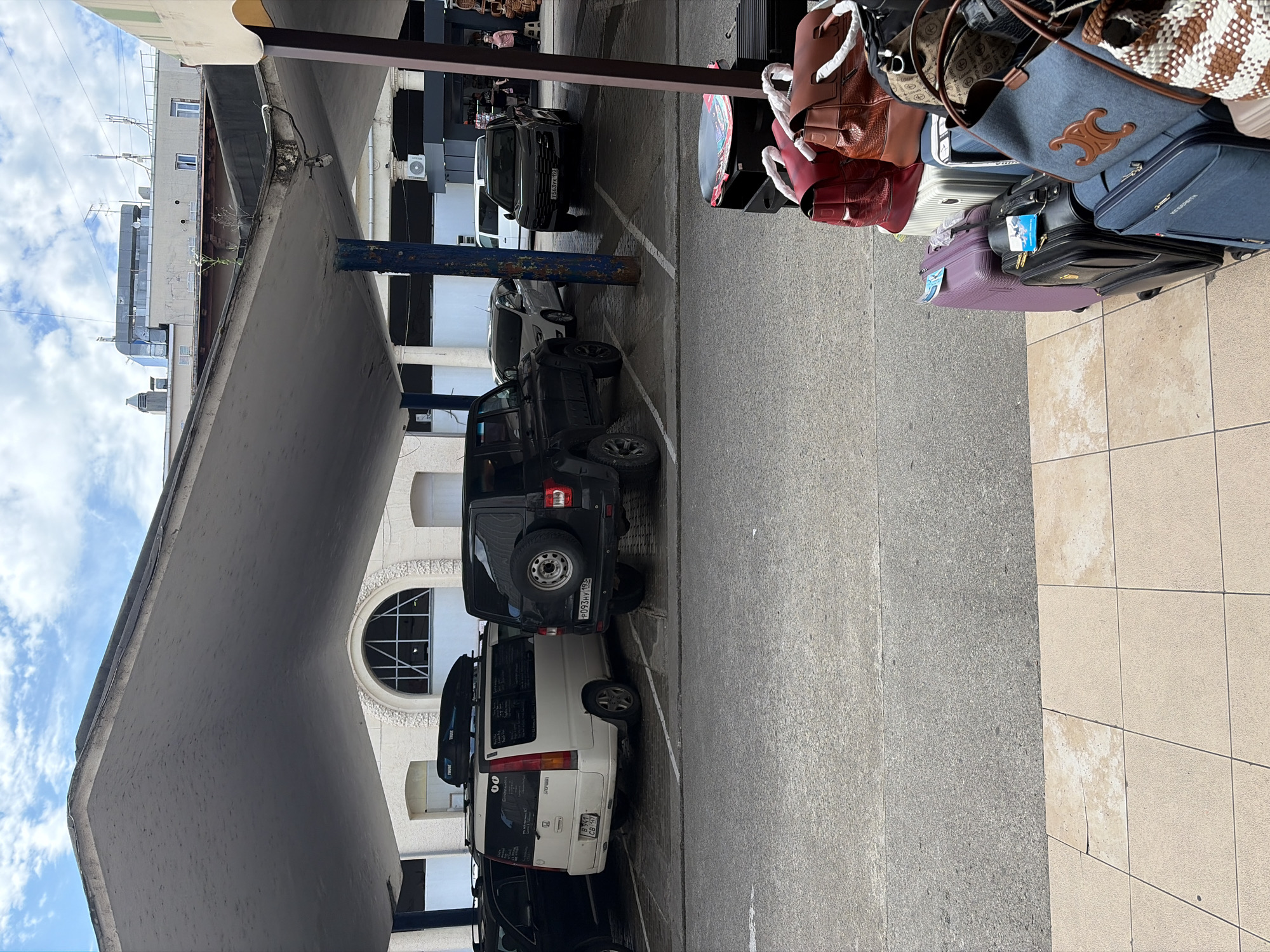 The image shows a parking lot under a large, curved, concrete canopy. The canopy is supported by thick, blue-painted columns. In the background, there is a building with an arched window. Several cars are parked in the lot, including a white van with a roof rack, and two dark SUVs. In the foreground, a stack of luggage is visible, including suitcases and bags in various colors and materials. The ground is paved with asphalt and tiled surfaces. The sky is visible overhead, with scattered clouds. The scene appears to be in a public area, possibly near a transport hub or market in Sochi, Russia. There are no people clearly visible in the image.