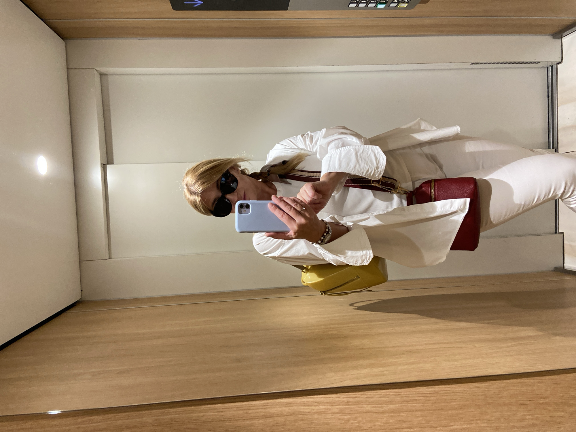 The original image is rotated 90 degrees clockwise. Described as if correctly oriented, the media file captures a woman taking a mirror selfie within what appears to be an elevator or a mirrored hallway.
**Subjects:** The primary subject is a woman with blonde hair, wearing large dark sunglasses, a white long-sleeved shirt or dress, and light-colored pants. She is holding a light blue smartphone in her right hand, positioned to take the photo. A red handbag with a distinctive striped (burgundy and white) strap is slung across her body, and a yellow handbag rests on the floor next to her reflected image. She wears a ring on her right hand and bracelets on her left wrist.
**Scene/Setting:** The setting is an indoor space characterized by extensive mirroring. The walls appear light beige or off-white, and the floor and ceiling are finished with warm-toned wooden panels. The entire scene is a reflection of the woman and her surroundings.
**Activity or Event:** The woman is actively taking a mirror selfie with her phone.
**Notable Details:** The lighting is artificial, suggesting an indoor setting. In the upper part of the reflected image, above the woman's head, an elevator control panel is visible on the wooden ceiling.
**Visible Text:** On the elevator control panel, a blue arrow pointing left is visible (`<-`), along with several small, illuminated square buttons.
The environment appears modern and well-lit, consistent with an upscale interior in a location like Budva, Montenegro.