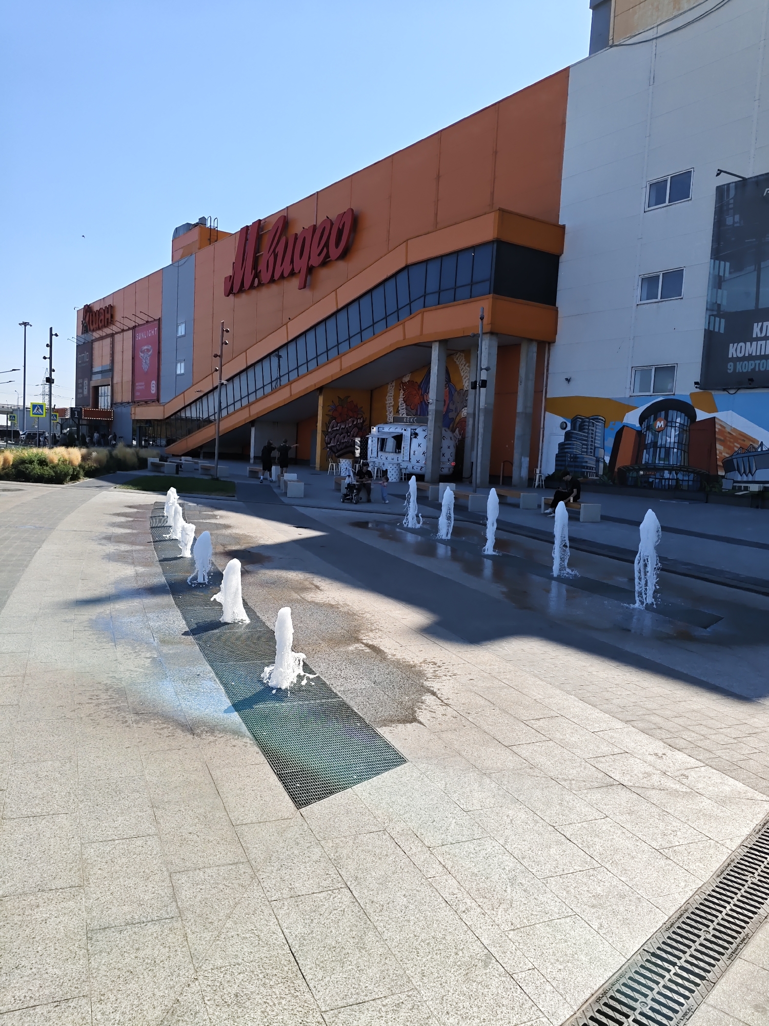 The image depicts the exterior of a M.Video store in Rostov-on-Don, Russia, under a clear blue sky. In the foreground, a row of modern fountains is active, with water jets spraying upwards from grates embedded in the paved plaza. The plaza is constructed of large, light-colored granite tiles. To the right, a drainage grate runs along the edge of the plaza. In the background, the M.Video building is prominent, featuring a large orange facade with a distinctive sloped, dark-windowed section. Several signs are visible on the building, including the M.Video logo in red script and advertisements for other stores like "Sunlight." A white food truck with Cyrillic text is parked near the entrance. Several people are visible in the scene, some walking, others sitting on benches, and one person is in a wheelchair. The environment appears to be an outdoor commercial area with some landscaping, including dry grasses and bushes to the left. The overall impression is a bright, sunny day in an urban commercial district.