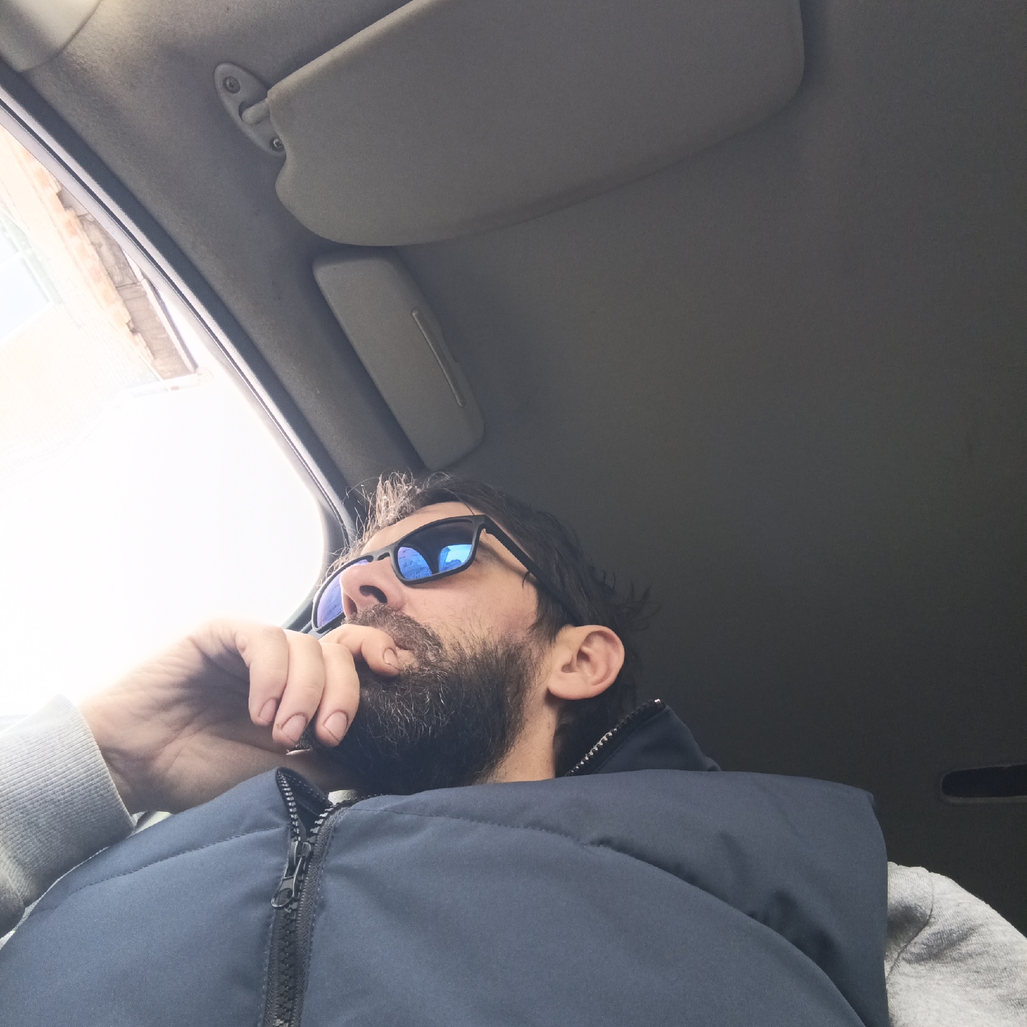 A man is sitting in a car. He is wearing sunglasses and a blue vest over a grey sweatshirt. He is touching his beard with his right hand, and appears to be looking out of the car window on the left side of the frame. The interior of the car is visible, including the ceiling and sun visor. The location is undetermined. There is no verifiable evidence in the image that places the location in the city of Chaikovsky, Russia.