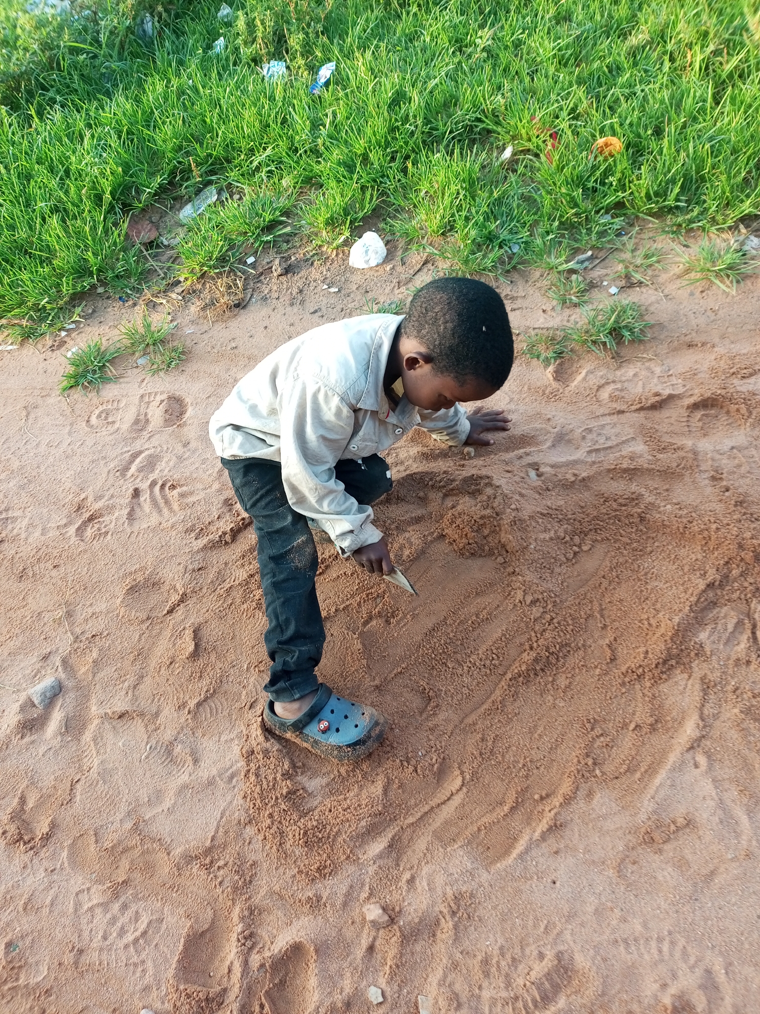 A young Black boy, likely between five and eight years old, is crouched down on reddish-brown sand, digging with a small shard of wood or plastic. He is wearing a light-colored, long-sleeved shirt, dark trousers, and a blue Croc-style shoe with a red cartoon face design on it. His hair is short and dark. The ground is composed of sand, with scattered footprints and some small pebbles. In the upper portion of the image, there is a strip of vibrant green grass, with some scattered litter, including plastic bags and wrappers. The scene appears to be an outdoor, unpaved area, possibly a roadside or vacant lot, within the city of Gombe, Nigeria. The lighting suggests it is daytime, with sunlight casting shadows.