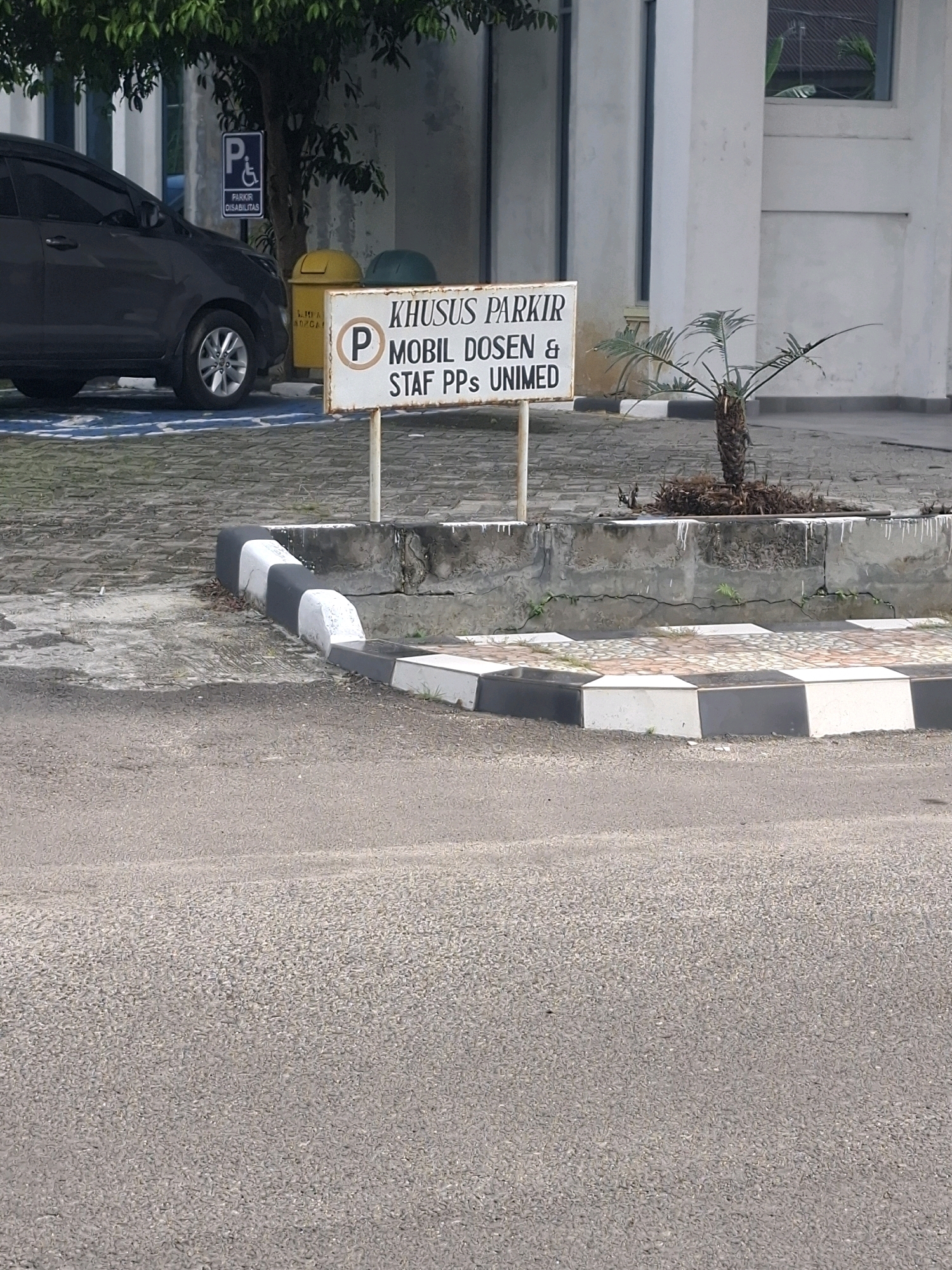 The image shows a parking lot area at the Politeknik (Polytechnic) Negeri Medan (Polmed) in Medan Estate, Indonesia. In the foreground, a paved road is visible. Behind it, there is a raised concrete planter box with a small palm tree. A sidewalk with black and white striped edging runs along the planter box. A white sign with black lettering reads "KHUSUS PARKIR MOBIL DOSEN & STAF PPs UNIMED," indicating that this parking area is reserved for lecturers and administrative staff of the Postgraduate Program at UNIMED (Universitas Negeri Medan). To the left of the sign, a dark gray car is parked in a designated parking spot marked with blue lines. Further left, a blue sign with a white "P" and a wheelchair symbol indicates a parking spot for persons with disabilities. Two trash bins, one yellow and one green, are situated behind the car. In the background, a modern building with white walls and windows is visible. The scene appears to be during daylight hours.