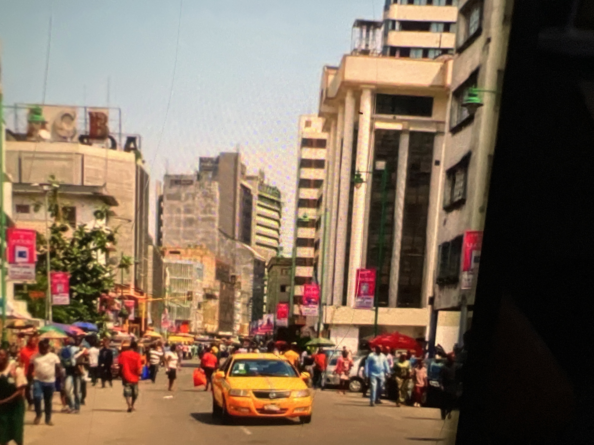 The image captures a busy street scene in Lagos, Nigeria, likely in a commercial or business district. The architecture features tall, modern buildings, some with distinct facades and repeating window patterns, interspersed with older structures. Several buildings display prominent signage, including a visible "C" and "B" on a rooftop in the upper left portion of the image, suggesting commercial establishments.
The street is populated with numerous people, a mix of pedestrians and vehicles. Pedestrians are engaged in various activities, including walking, socializing, and potentially shopping, with some individuals clustered around stalls or umbrellas, indicative of a marketplace. A bright yellow taxi is prominently featured in the foreground, suggesting its active use for transportation. Other vehicles are visible in the background.
Decorative banners and signs, many in red, are hung along the street, contributing to a vibrant and perhaps festive atmosphere. The overall impression is one of a bustling urban environment, characteristic of a major city like Lagos, with a blend of commercial activity, diverse populations, and dynamic street life. The lighting suggests it is daytime, with a clear, bright sky.