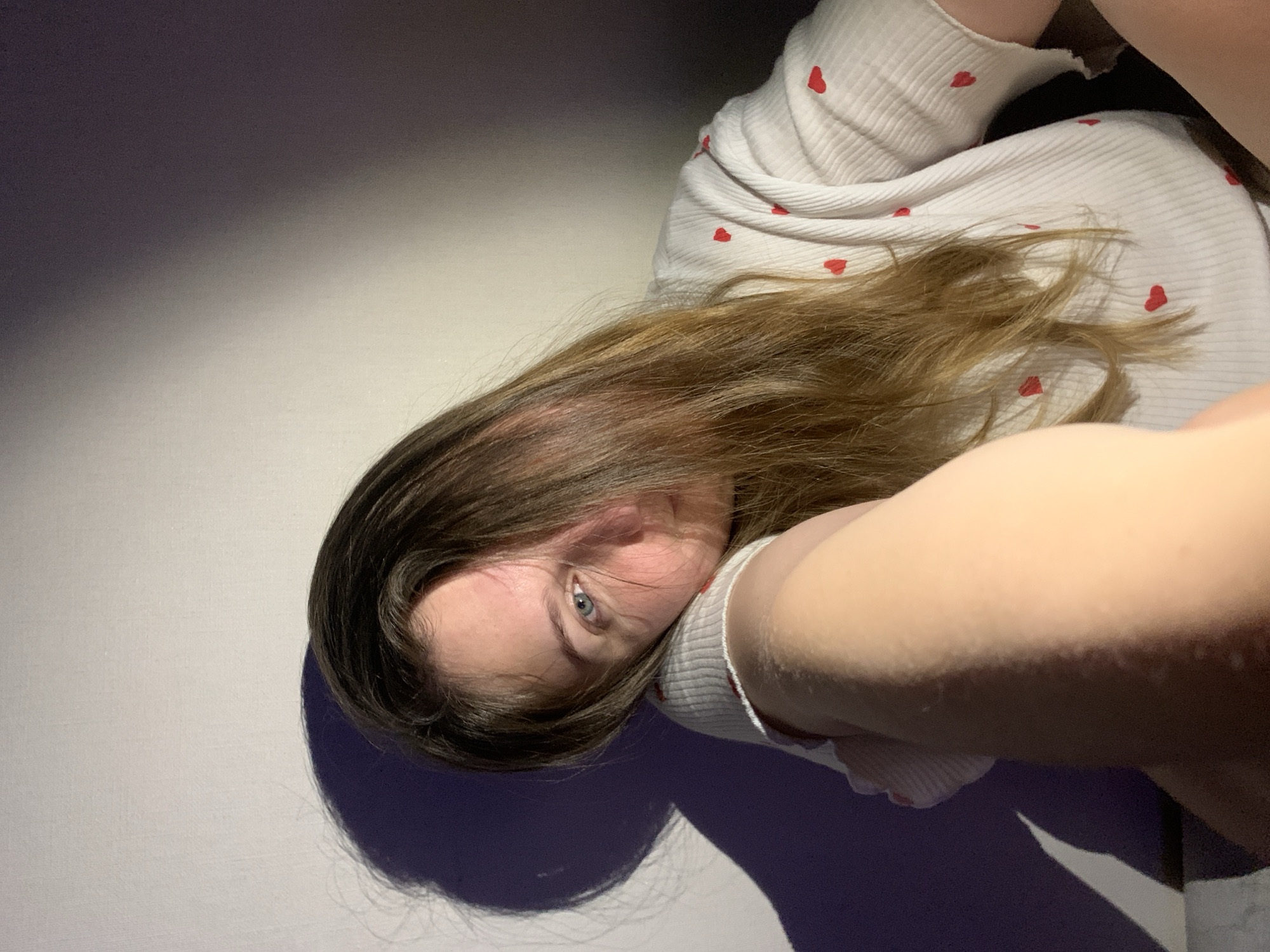 The image captures a close-up, angled view of a young individual, likely female, lying indoors, possibly in Chaikovsky, Russia. The subject has fair skin, brown hair, and a visible blue eye, looking directly into the camera with a neutral expression. She is wearing a white ribbed top patterned with numerous small red hearts.
The perspective suggests the person is lying on her side or back, with her head partially supported by her arm or another surface, while looking upwards at the camera. A portion of her arm is prominently in the foreground, out of focus. The background consists of plain, light-colored surfaces transitioning into darker, shadowed areas of purple and gray, indicative of artificial lighting casting strong shadows. These shadows are particularly noticeable around her head and body. The setting appears to be a generic indoor space, such as a floor or bed, offering no specific location cues for Chaikovsky, Russia, other than the provided context. The activity depicted is a posed photograph, possibly a selfie. There is no visible text, and the time of day is indeterminate due to the artificial lighting.