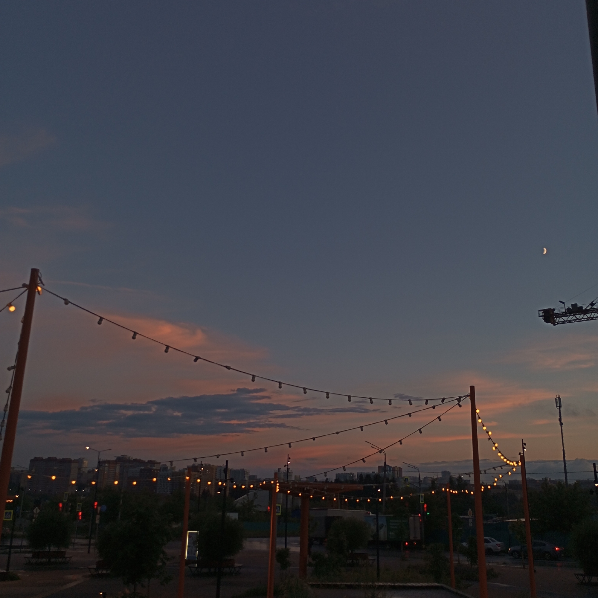 A twilight scene in Pervomaysky, Russia, captures the sky with a gradient of deep blue and streaks of pale orange and pink clouds. A crescent moon is visible in the upper right quadrant. In the foreground, strings of illuminated bulbs stretch across the frame, supported by orange poles. These lights cast a warm glow against the darkening sky. Below the lights, a city skyline with apartment buildings is visible, punctuated by streetlights and traffic signals. Trees and vehicles are also present, suggesting an urban environment. A construction crane is visible in the far right of the image. The overall impression is one of a city transitioning from day to night, with the artificial lights of the city and decorative bulbs mingling with the natural light of the evening sky.
