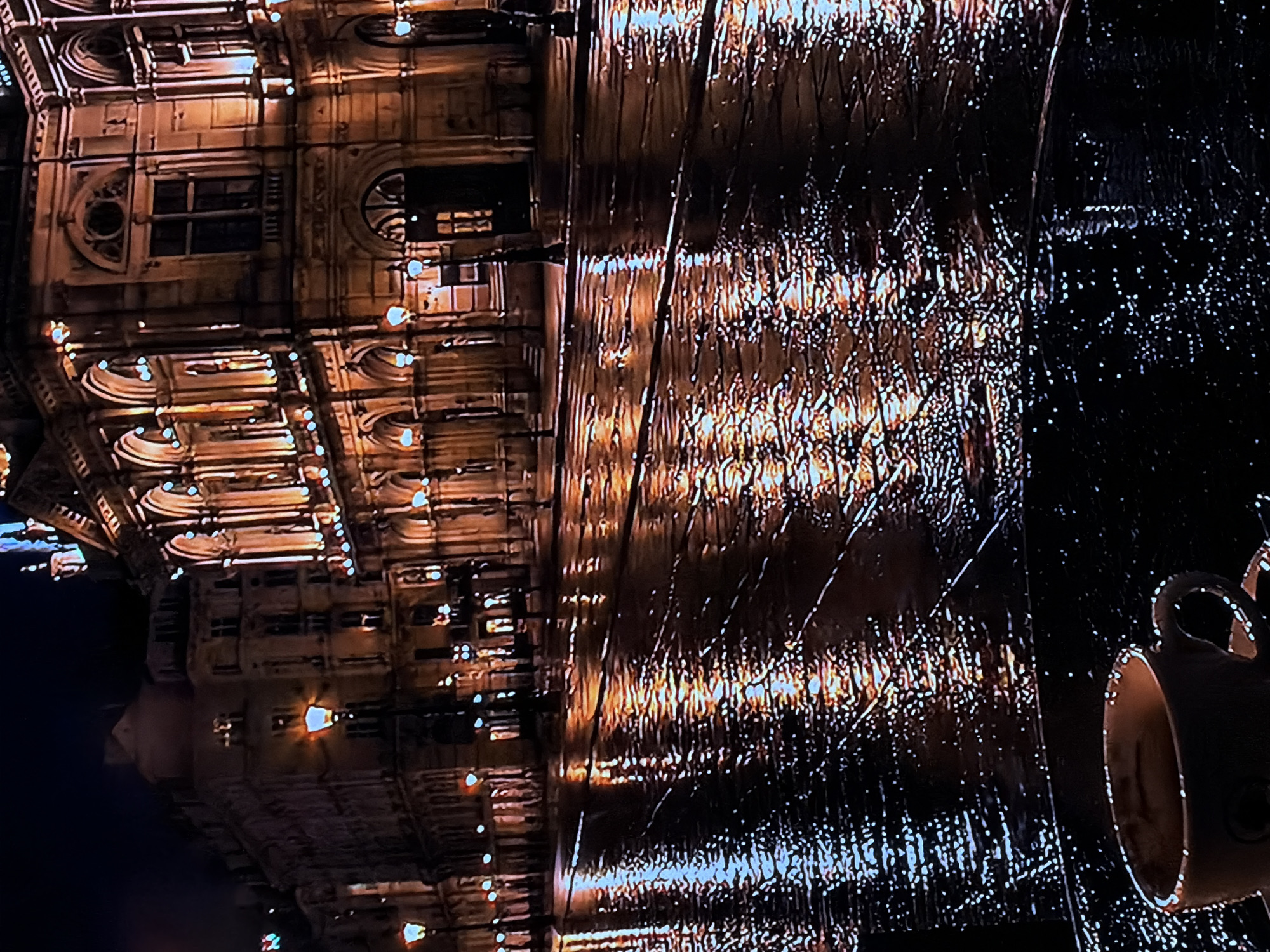 The media file displays a nighttime scene, rotated 90 degrees clockwise, showing an illuminated ornate building reflected on a wet, dark surface. The building, positioned on the left side (if rotated upright), is multi-storied with intricate architectural details including arched windows, circular accents, and columns. It is brightly lit by numerous warm, golden lights that highlight its features and cast a glow, suggesting a historic or classical architectural style.
The prominent reflection occupies the right side of the image, appearing as elongated streaks and scattered points of light on what seems to be a wet street, a large puddle, or a highly reflective surface like a wet tabletop. The texture of the reflection implies recent rain or moisture.
On the far right, partially cropped, a ceramic mug with a handle is visible, also reflecting some of the ambient light. Its interior appears light-colored. No people are present in the scene. The overall impression is one of a quiet, illuminated urban night, possibly in Sofiivska Borshchahivka, Ukraine, given the context. There is no visible text in the image.