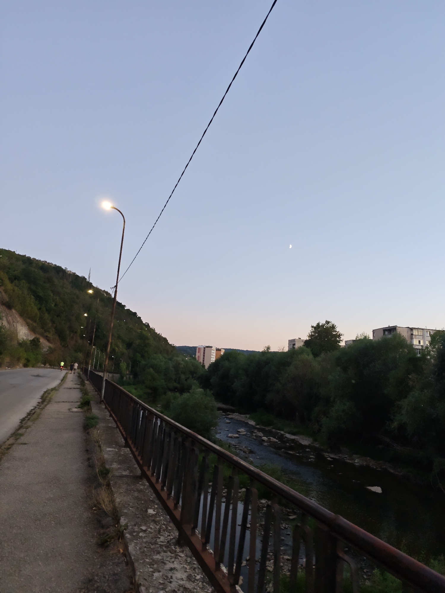 The image captures a twilight scene in Lovech, Bulgaria, looking east from a bridge. A paved pedestrian walkway runs alongside a road, bordered by a rusty metal railing. Beyond the railing, a narrow, rocky river flows through a densely wooded area. On the left, a steep, tree-covered hillside rises from the road, with a few streetlights illuminating the walkway. In the distance, apartment buildings are visible amongst the trees, and a sliver of the moon hangs in the pale blue sky. Two figures, likely pedestrians, are walking away from the camera on the walkway.