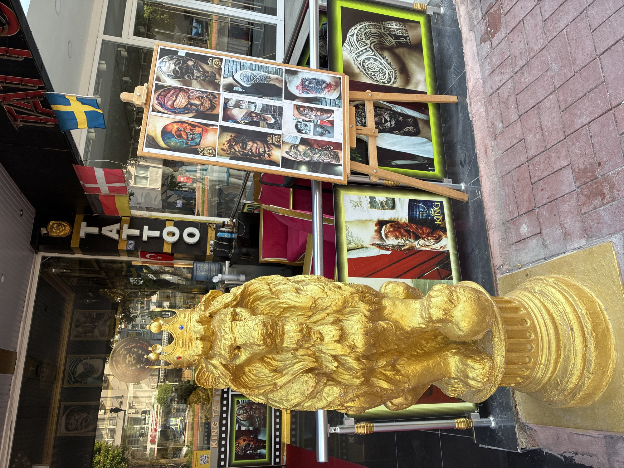 The image shows the exterior of a tattoo shop called "King Tattoo" in Alanya, Turkey. A large, golden lion statue wearing a crown stands in front of the shop. An easel displays a collage of tattoo designs, primarily featuring skulls and intricate patterns on skin. Flags of Germany, Denmark, and Sweden are visible, suggesting an international clientele or the owner's heritage. The shop's entrance is visible, with windows reflecting the street scene and surrounding buildings. The shop appears to be located on a pedestrian-friendly street with brick paving.