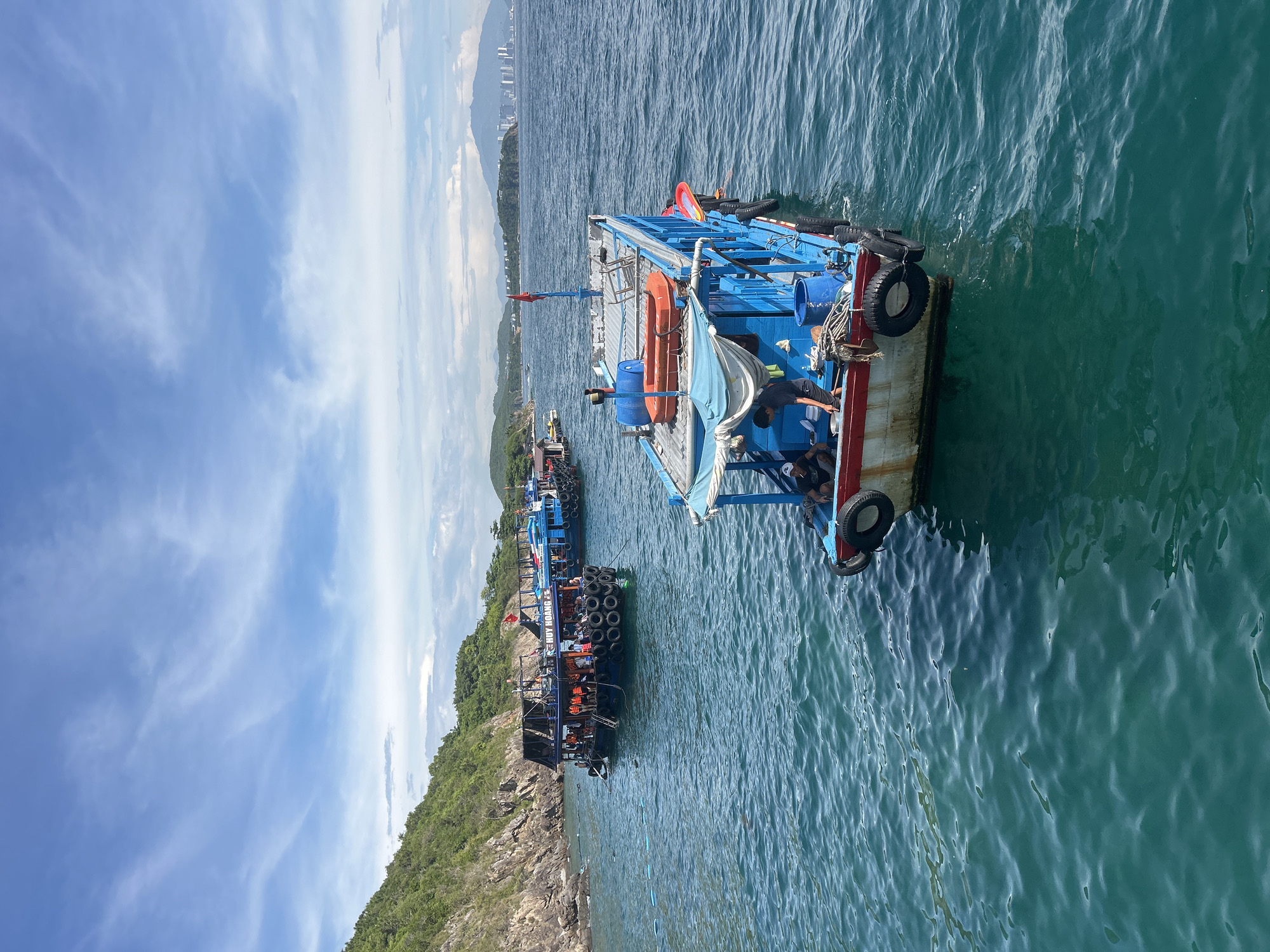 The image depicts a scenic view of the coast of Nha Trang, Vietnam, with several boats docked in the turquoise waters. In the foreground, a blue and white boat is anchored, with two individuals on board. The boat features a red trim and is equipped with life preservers, suggesting it may be used for tourist excursions. In the midground, a larger dark-colored boat, possibly a ferry, is filled with passengers wearing orange life vests. This boat displays the name "HUY HOANG" on its side and appears to be navigating through a bay. Further in the distance, additional boats are visible, and the coastline is characterized by lush green hills. The sky is partly cloudy, with a mix of blue sky and white wispy clouds. The cityscape of Nha Trang can be observed in the far background, with buildings lining the coast and mountains forming a backdrop.