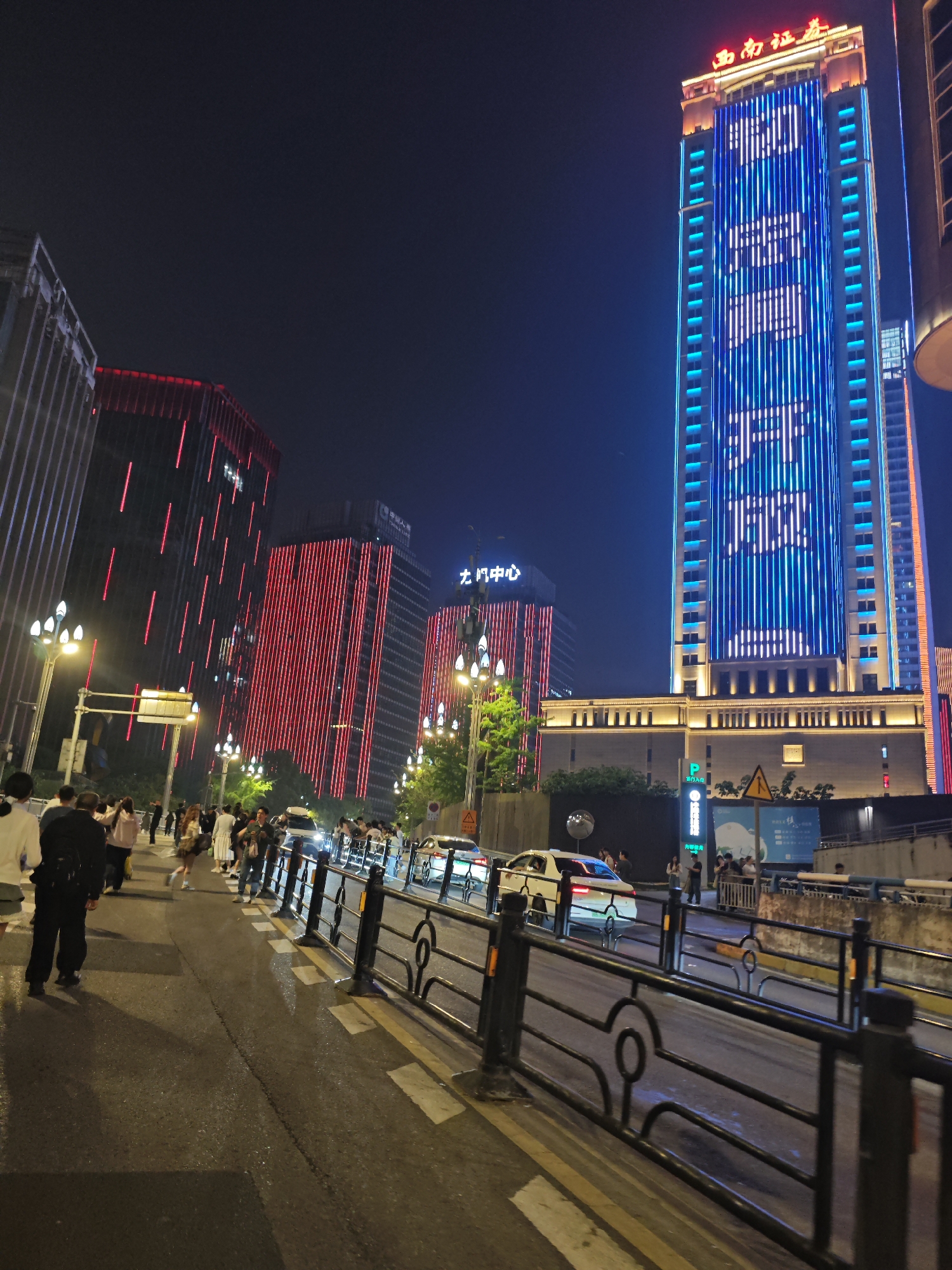 Captured at night in 江北城街道, China, the image shows a bustling cityscape featuring a tall, illuminated skyscraper. This building is decorated with blue lights outlining its structure and displays a vertically oriented text or design in white lights down the center. Adjacent to the skyscraper are other buildings lit in red.
A pedestrian walkway is lined with a black metal fence that separates it from the street. People are walking and standing on the walkway. On the street, vehicles, including at least two white cars, are moving. A sign is visible near the street, and street lights illuminate the scene.