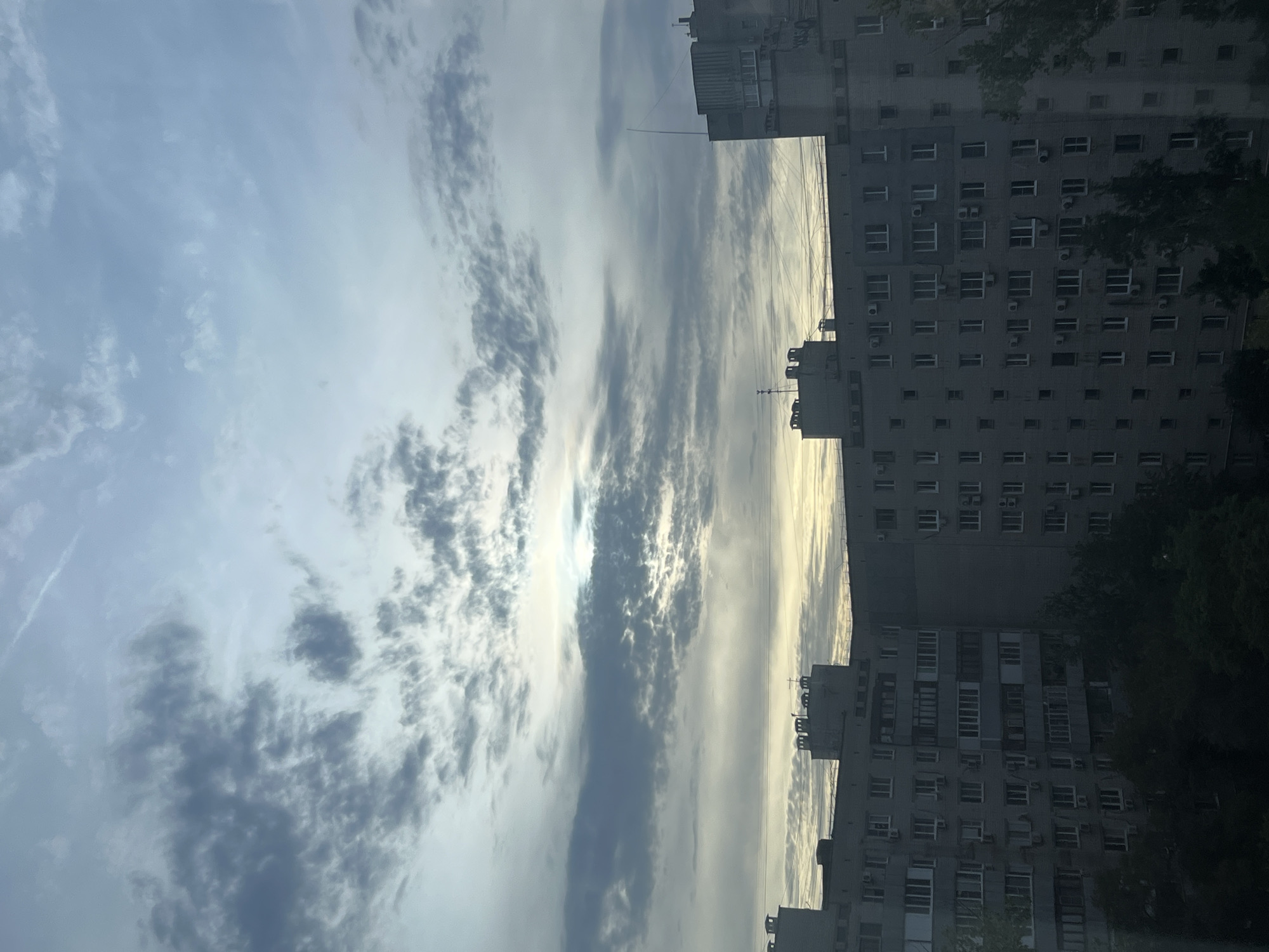 The image captures a view of Soviet-era apartment buildings under a cloudy sky in Volgograd, Russia. The buildings are tall, monolithic structures with numerous windows and balconies, characteristic of this architectural style. The sky is filled with a mix of dark and light gray clouds, with hints of yellow near the horizon, suggesting either sunrise or sunset. Some antennas and ventilation units are visible on the rooftops of the buildings. Trees with dark green foliage are sparsely scattered in the foreground, obscuring the lower portions of the apartment blocks. There are no people visible in the image. The overall impression is of a typical urban residential area in a cloudy, diffused light.