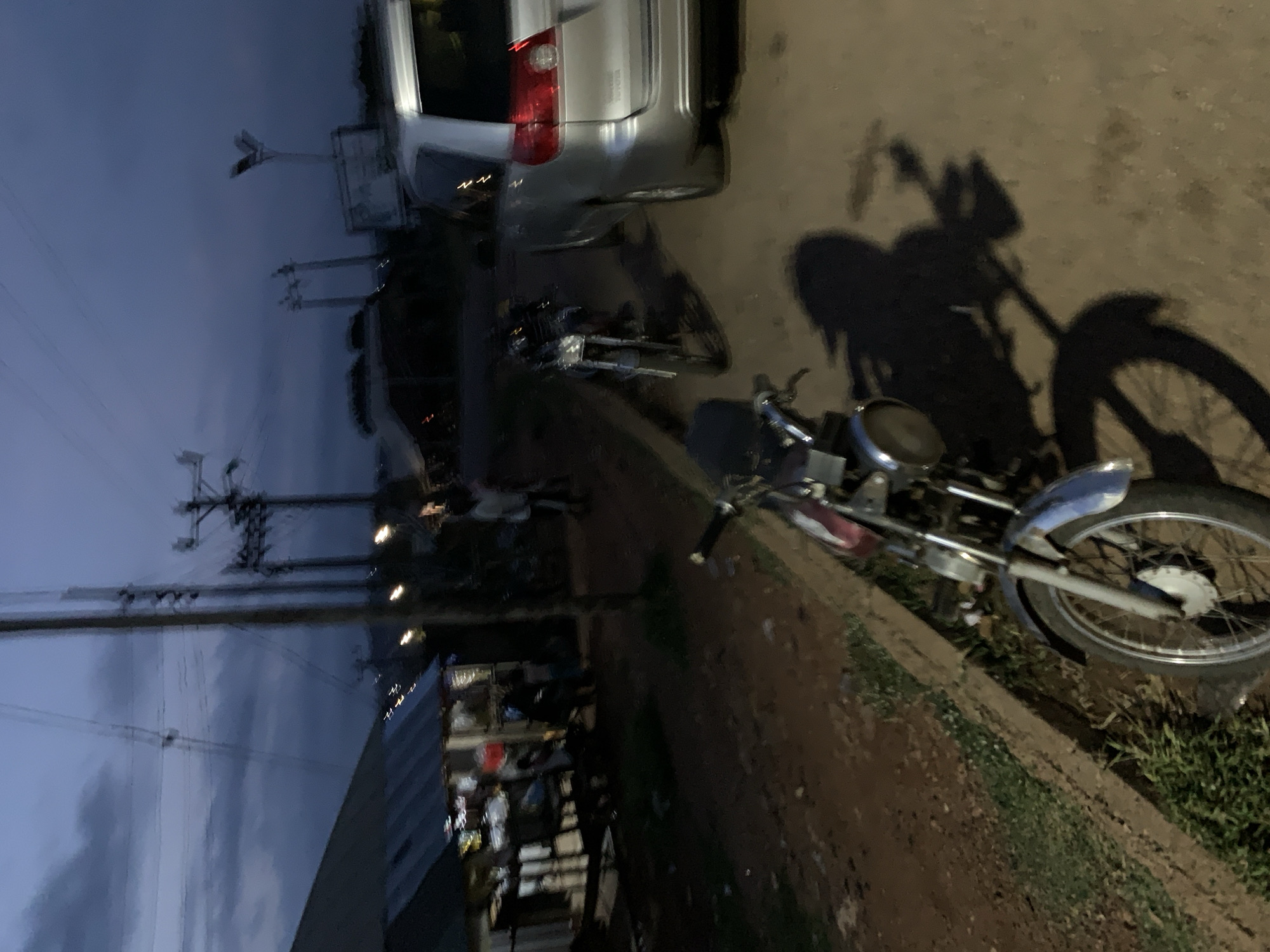 The image depicts a street scene at dusk in Kabba, Nigeria. In the foreground, a motorcycle is parked facing the right, with its front wheel and handlebars visible. The motorcycle has a silver fender and a round headlight. To the right of the motorcycle is a shadow cast by the motorcycle and potentially another object. Further into the scene, a silver minivan is parked, its rear section illuminated by ambient light. Behind the minivan, another motorcycle is visible, parked parallel to the road. On the left side of the image, a low-slung structure with a corrugated metal roof stands, possibly a market stall or shop, with several people standing or sitting around it. The ground appears to be dirt and gravel. In the background, there are utility poles with electrical wires stretching across the frame, and a cloudy sky. The overall atmosphere is dim, suggesting either early morning or late evening.