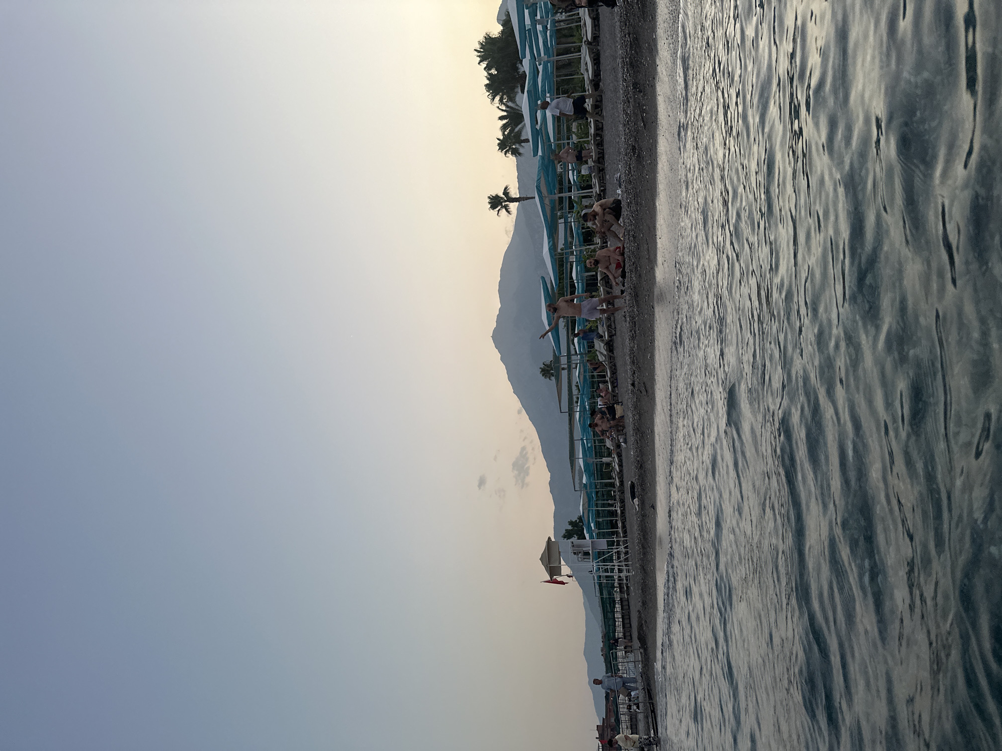 The image depicts a serene beach scene in Kemer, Turkey, during the late afternoon or early evening. The foreground is dominated by the gentle waves of the Mediterranean Sea, with their surface reflecting the soft, diffused light of the setting sun. Beyond the water lies a pebble beach, sparsely populated with a few individuals and groups of people.
In the mid-ground, a row of blue and white striped beach umbrellas provides shade over loungers, suggesting a resort or public beach area. A lifeguard tower, topped with a red flag, stands prominently near the center of the frame. Several people are visible scattered across the beach, some sitting, others standing, and one man is raising his arms as if in greeting or admiration. Palm trees are interspersed with the umbrellas, adding to the tropical ambiance.
The background is characterized by a majestic mountain range, its slopes softened by the distance and the dusky light. The sky above is a pale blue, transitioning to a warmer, lighter hue near the horizon, indicating the approaching end of the day. The overall impression is one of tranquility and relaxation, a typical scene at a popular coastal destination in Turkey.