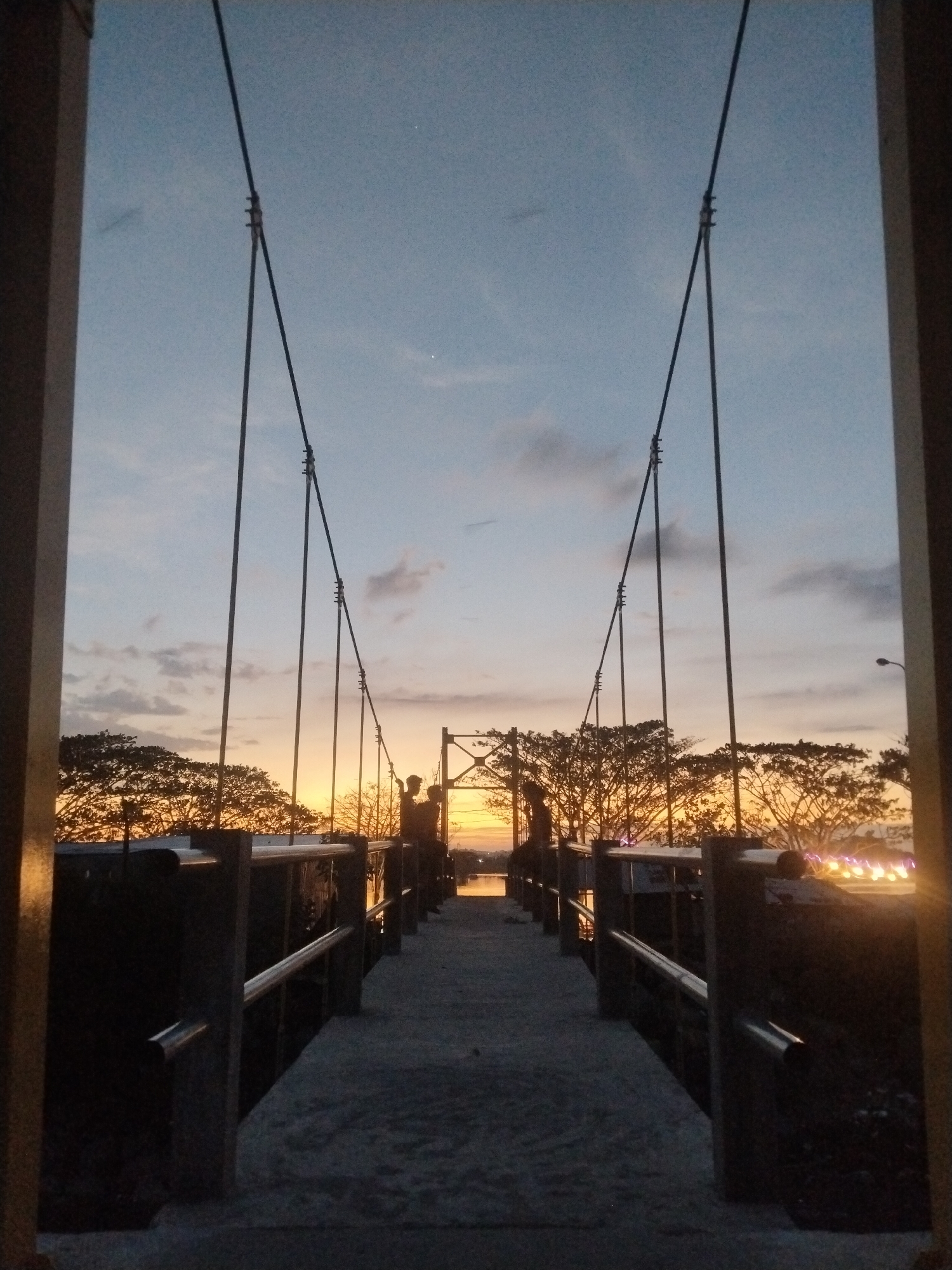 The image, taken in Lhokseumawe, Indonesia, shows a suspension bridge at dusk. The bridge's concrete walkway is flanked by metal railings and supported by cables extending upwards. Silhouetted figures of two individuals stand on the bridge's far end, possibly interacting. Trees are visible beyond the bridge, their silhouettes highlighted by the setting sun. Light fixtures illuminate the right side of the bridge. The image is framed by vertical posts, suggesting it was taken from a vantage point on land. The sky is a mix of blue and orange hues, indicating a transition between day and night.
