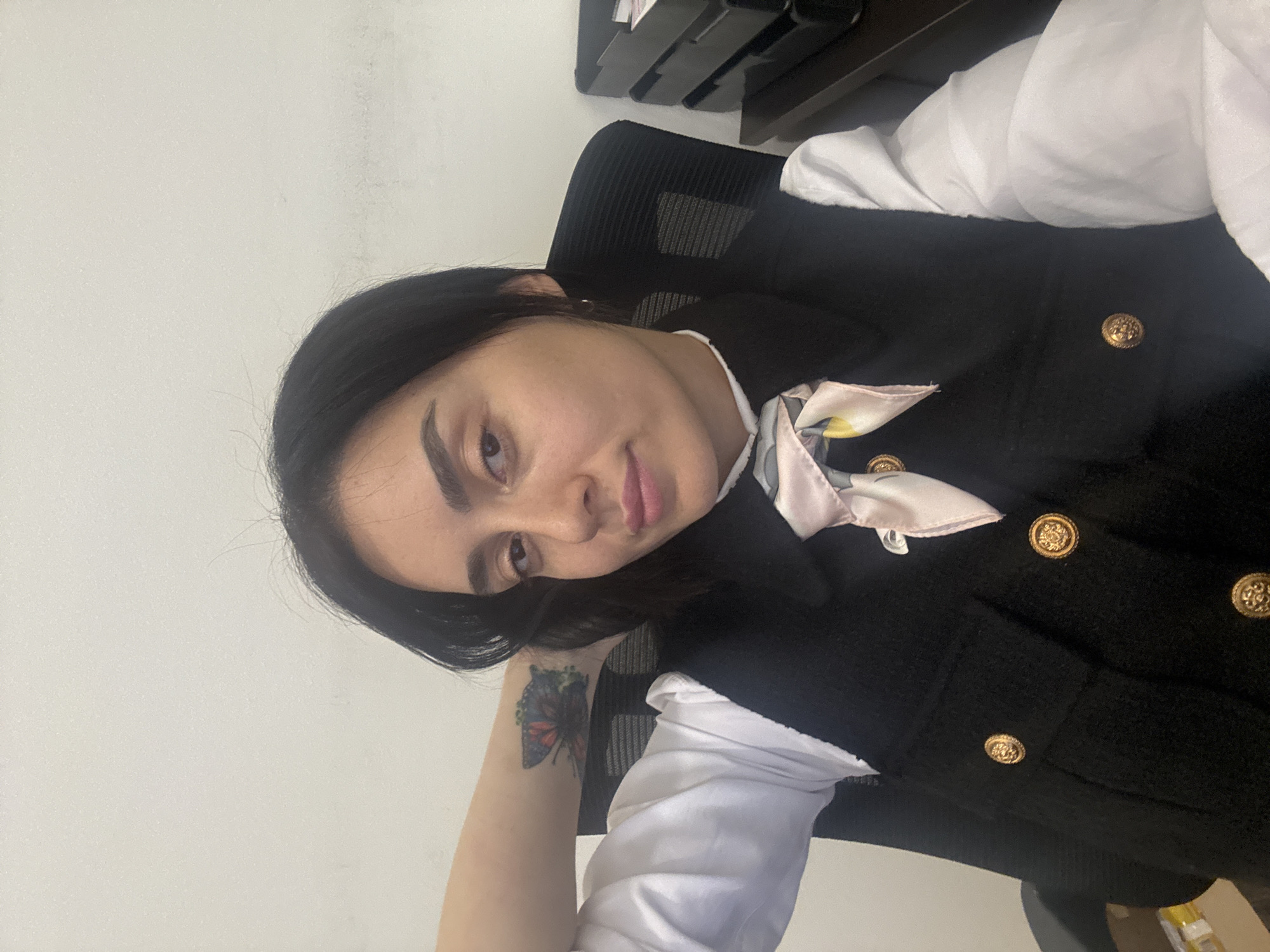 A young woman with dark hair and dark eyebrows is the central subject of this indoor selfie. She is wearing a white long-sleeved collared shirt under a black textured vest or jacket adorned with gold, ornate buttons, two of which are clearly visible. A light pink or cream-colored silk scarf with a subtle floral or abstract pattern is tied around her neck. On her left forearm, a colorful butterfly tattoo is visible, featuring shades of blue, red, and green. She is seated in a black office chair with a mesh backrest.
The woman is taking a close-up selfie, looking directly into the camera with a soft, pleasant smile and slightly parted lips. Her head is tilted slightly. The setting appears to be an office or workspace, indicated by the office chair and a dark shelving unit with document trays visible in the background, against a plain light-colored wall. The lighting is bright and even, suggesting daytime or well-lit indoor conditions. No specific time of day or weather is discernible from the image itself. There is no visible text. The location context provided is Tbilisi, Georgia.