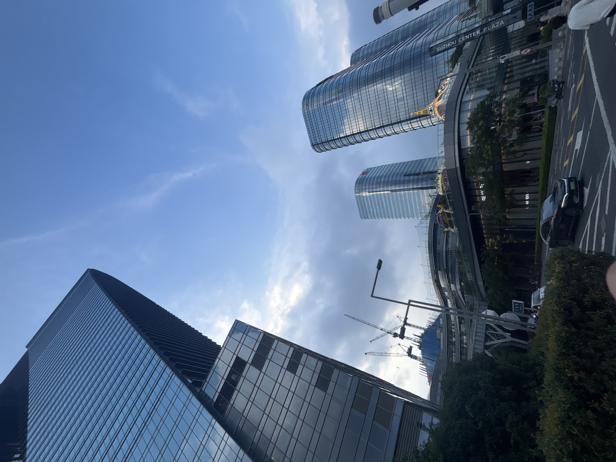 The image shows a street-level view of the Suzhou Center Plaza in Suzhou Industrial Park, China. The scene features modern architecture with several tall, glass-facade skyscrapers, some of which are curved. In the foreground, a street with lane markings is visible, with cars and a scooter driving on it. Lush green bushes and trees line the street and are integrated into the building's lower levels. Pedestrians are present on the sidewalk. Construction cranes are visible in the background, indicating ongoing development. The sky above is partly cloudy with a light blue hue. One building prominently displays the text "SUZHOU CENTER PLAZA" vertically.