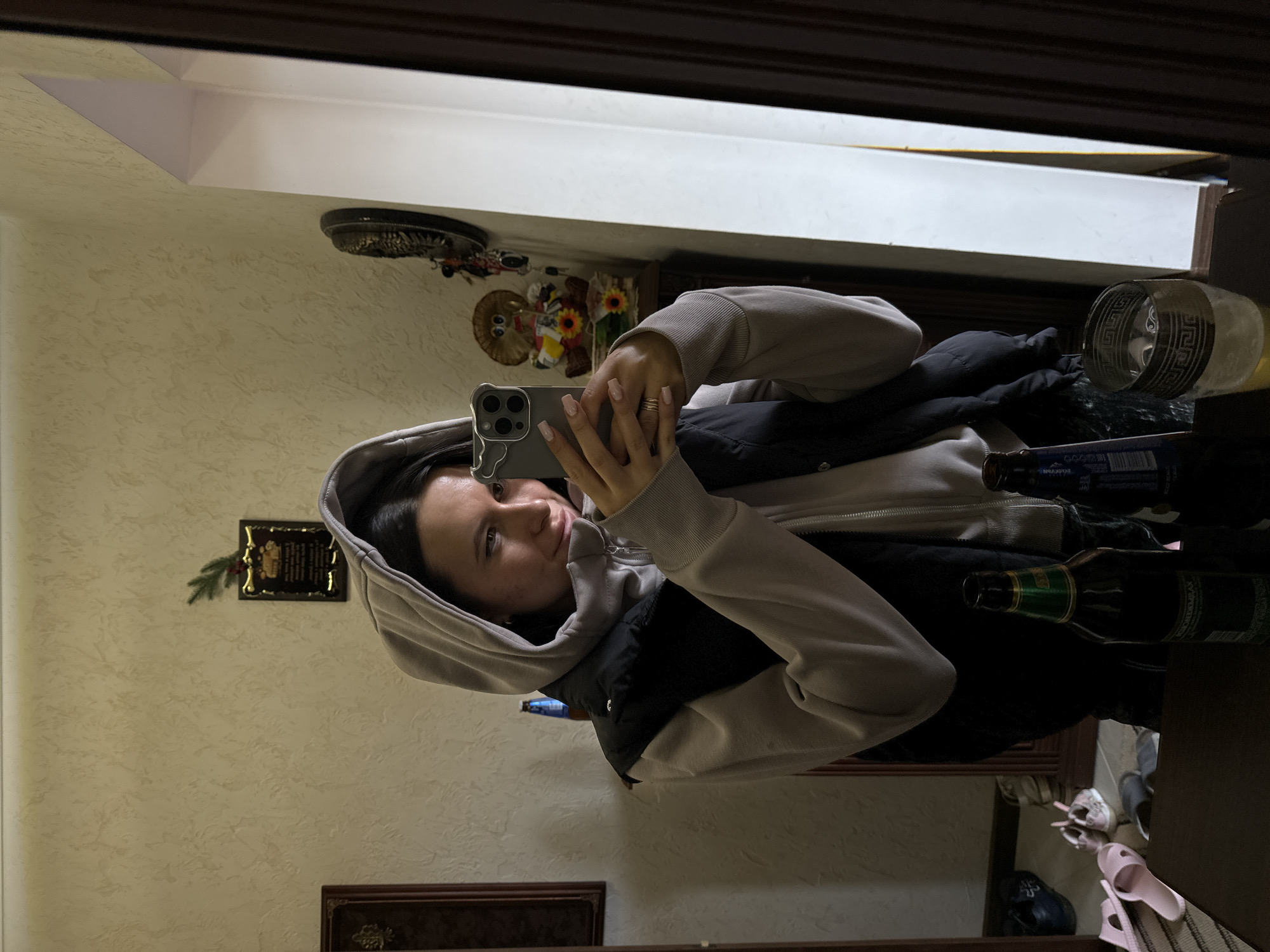A woman is taking a selfie in a mirror. She is wearing a gray hooded sweatshirt and a black vest. Her phone is gray with three lenses. There are bottles of beer, other objects, and various pairs of shoes in a cluttered space reflected in the mirror. On the wall behind her is a decorative wall plate. The location is likely in Vysokinichi, Russia.