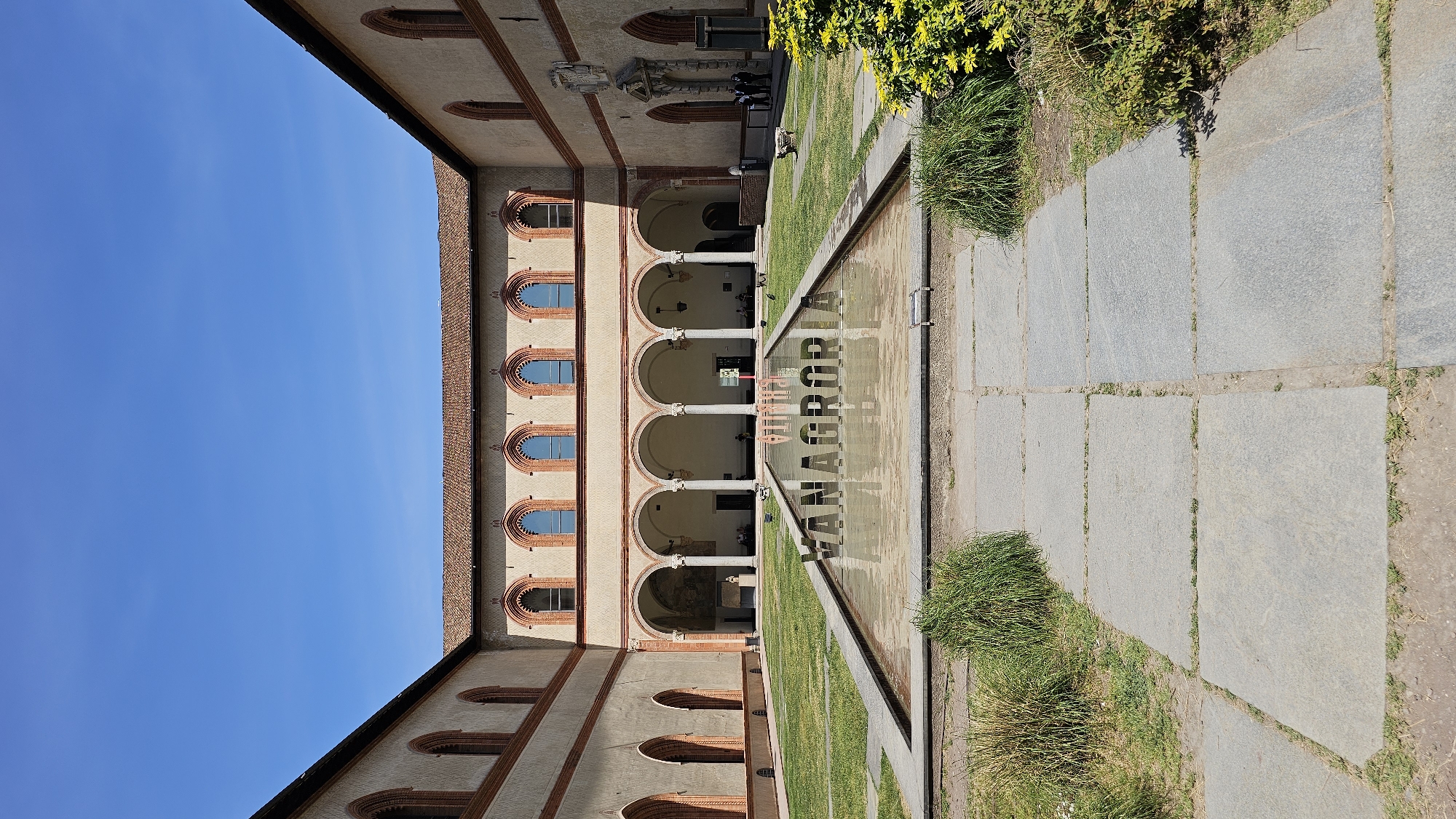This image depicts the courtyard of the Sforza Castle in Milan, Italy, on a sunny day. The courtyard is characterized by a long rectangular reflecting pool with the words "VANAGLORIA" visible in its reflection. The pool is flanked by stone pathways and patches of grass and vegetation. On either side of the courtyard stand the castle's architectural elements, featuring arcaded walkways with columns and rounded arch windows. The building's facade is composed of beige-colored stone with red brick detailing. People are visible in the background, underneath the arcade. The sky above is clear and blue.