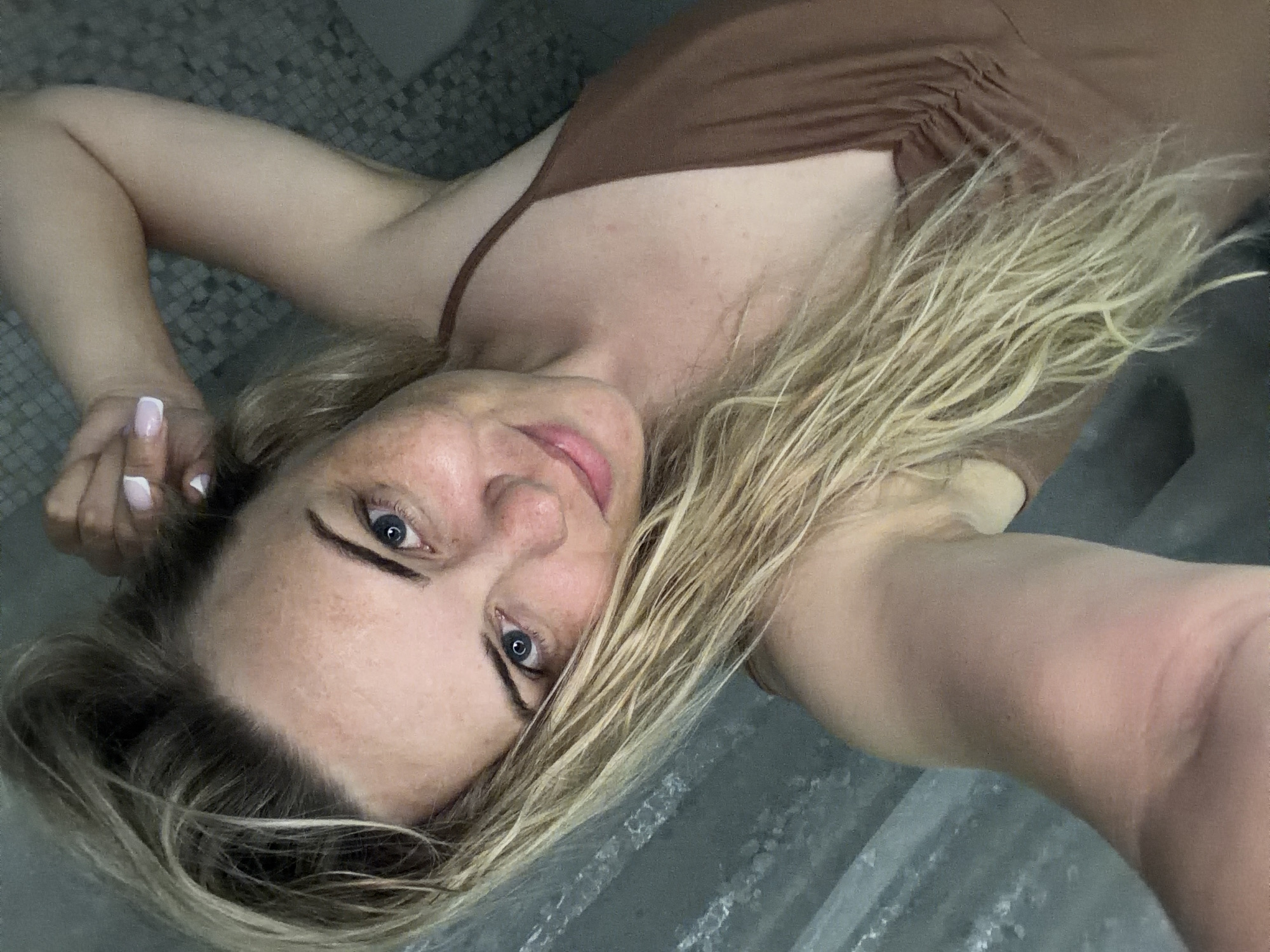 A selfie taken from a low angle of a fair-skinned woman with blue eyes and blonde hair, lying down on a light gray tile surface. She is wearing a brown tank top. Her right hand, visible in the upper left corner, has manicured nails with a white tip design. The background consists of the light gray tile surface with a slightly marbled or textured appearance. The location is not discernible as Votkinsk, Russia.