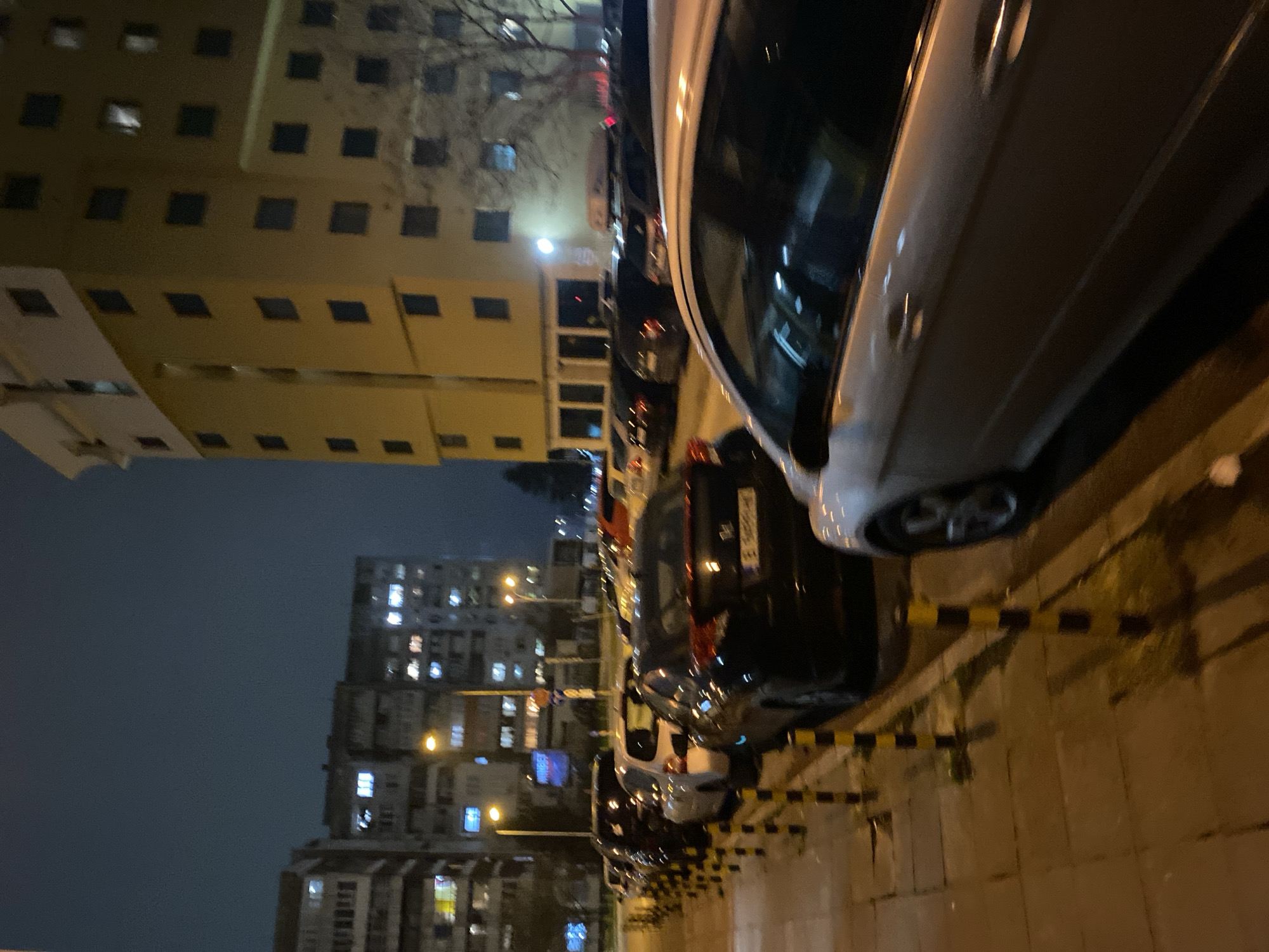 The image depicts an urban street scene at night in Varna, Bulgaria. The camera's perspective is angled upwards, showing tall apartment buildings with numerous illuminated windows on the left side. The buildings appear to be older, with a boxy architectural style. A streetlight illuminates the street.
Parked vehicles line the right side of the street, including a white car dominating the foreground. A black car is positioned behind the white car. Black and yellow striped bollards are visible along the curb, separating the sidewalk from the parked cars. The street surface is paved with stone. The overall atmosphere is dark, typical of a night scene.